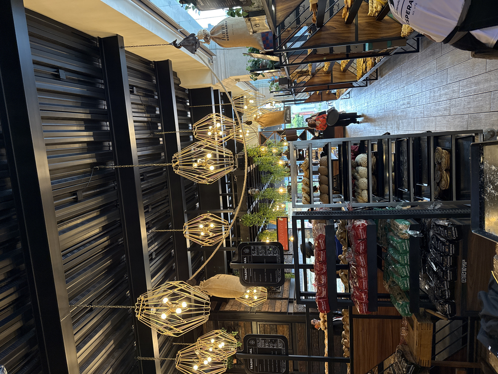 The image depicts the interior of a bakery named "Esperanza" located in Puebla, Mexico, as indicated by the "Esperanza MX. 1975" sign. The view is looking down a central aisle from front to back, with an array of geometrically shaped light fixtures suspended from the black metal ceiling supports with chains and rope. Sacks of flour hang above. On the right, shelves display a variety of baked goods including bread and pastries. A woman in an orange shirt holds a tray and stands near the back of the bakery, near the entrance. On the right side, shelves are stocked with packaged bread such as Integral and Centeno. A display with black menu boards showing prices for breads such as Campesino and Bolillo stands in the aisle. The floors are of light colored tile.