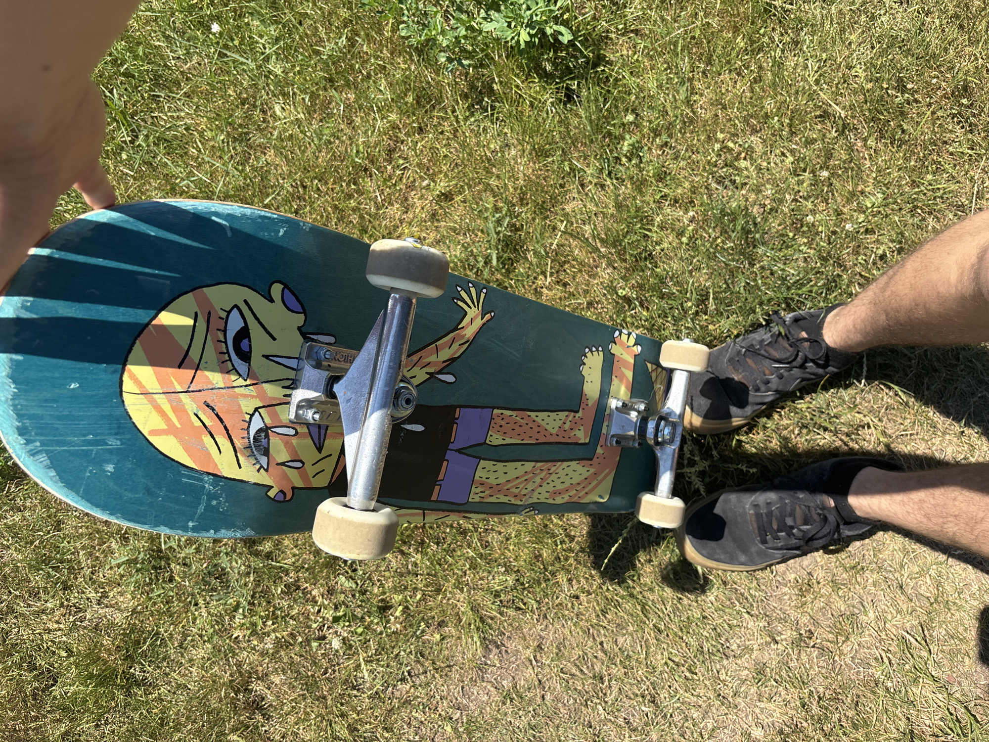 A person's legs and feet are visible in the lower part of the image, clad in black lace-up skate shoes. The person is standing on a grassy area, and a teal skateboard with a colorful, cartoonish graphic of a crying face and a body with pixelated shorts and patterned legs is positioned in front of them, angled slightly towards the camera. The skateboard is equipped with silver trucks and cream-colored wheels. A shadow from the skateboard and possibly a tree is cast on the grass. The scene appears to be outdoors on a sunny day.