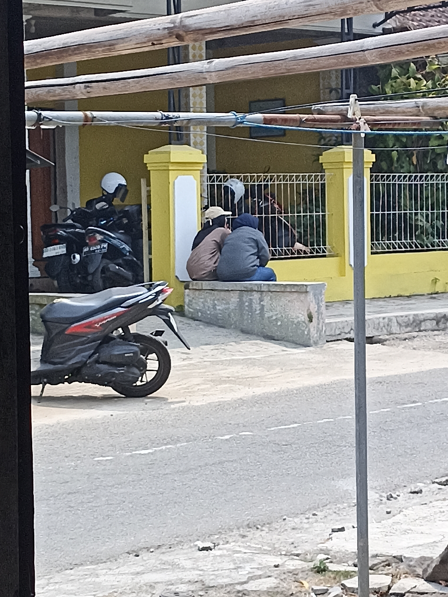 The image depicts a street scene in Pulisen, Indonesia, with a focus on a concrete low wall or ledge in the foreground. On this wall, three individuals are seated. Two of the individuals, wearing headscarves, are in close proximity, facing away from the camera and towards a yellow building with a white fence. The third individual, also wearing a headscarf, is seated to their left, also facing away from the camera. To the left of the wall, a dark-colored motorcycle is parked. In the background, the yellow building is visible, along with a white fence. The overall environment suggests an outdoor, urban setting.