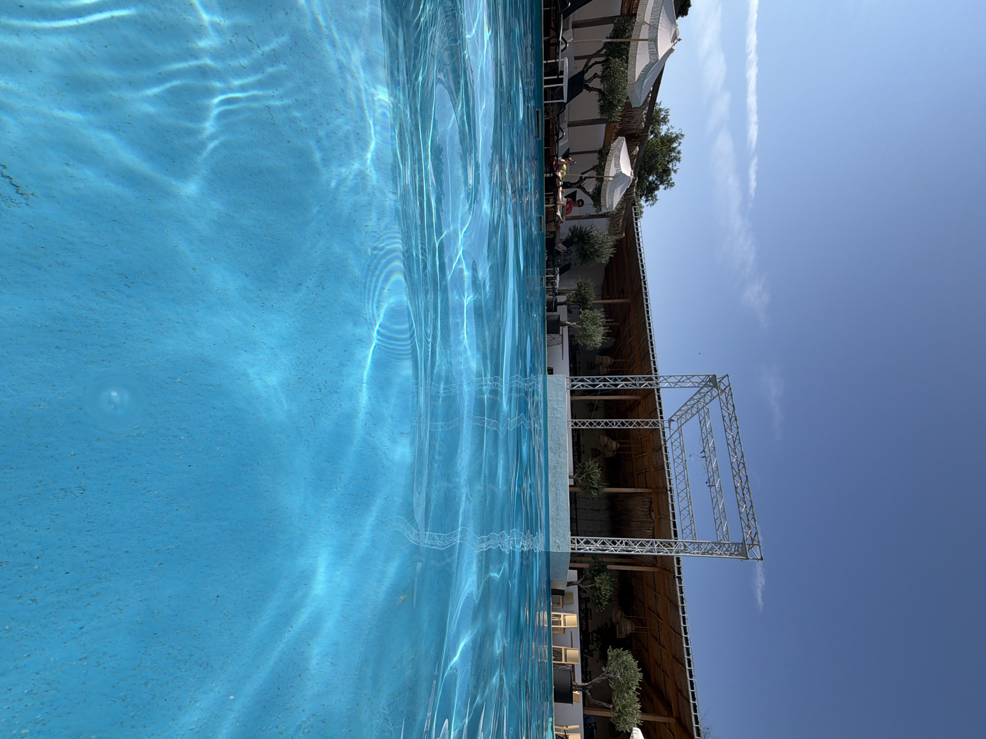The image depicts a clear blue swimming pool in the foreground, with sunlight creating rippling patterns on the water's surface. In the background, a covered outdoor area with a thatched roof is visible, supported by white columns and metal scaffolding. Several olive trees are planted around this area, and white umbrellas are set up over lounge chairs where two individuals are relaxing. The scene appears to be a resort or a poolside establishment. The sky above is a bright, cloudless blue. The location is the outdoor pool area of a resort in Burgas, Bulgaria.