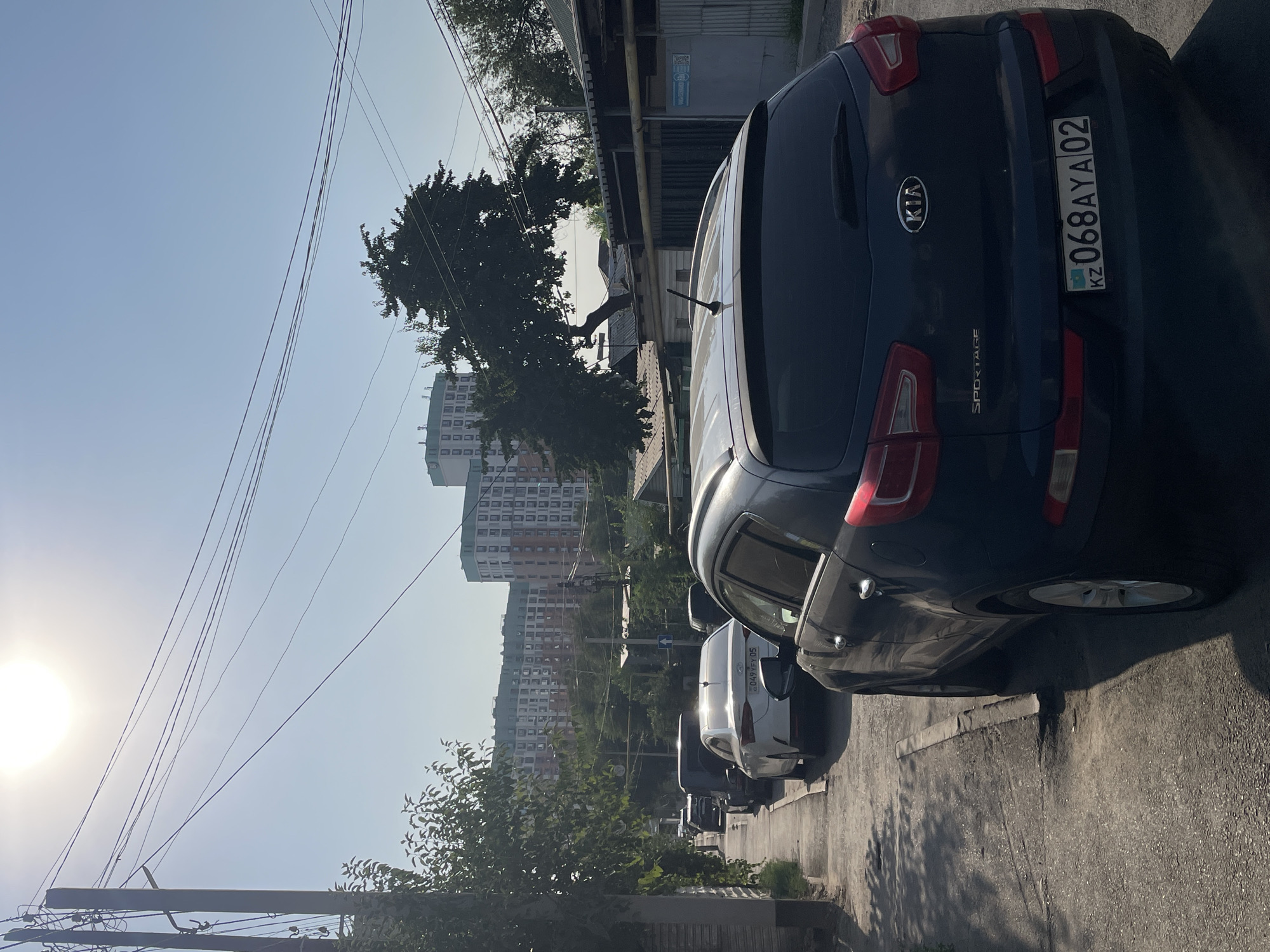 The image shows a street scene in Almaty, Kazakhstan, on a sunny day with a clear blue sky. Several cars are parked along the side of a paved road. In the foreground, a dark-colored Kia Sportage is prominently displayed, with its license plate reading "KZ 068AYA 02". Behind it, a white sedan is parked, followed by other vehicles. Tall, multi-story apartment buildings, likely in a residential area, dominate the background, with some trees and power lines visible. The overall impression is of a typical urban street in Almaty, with vehicles parked along the road and modern buildings in the distance.