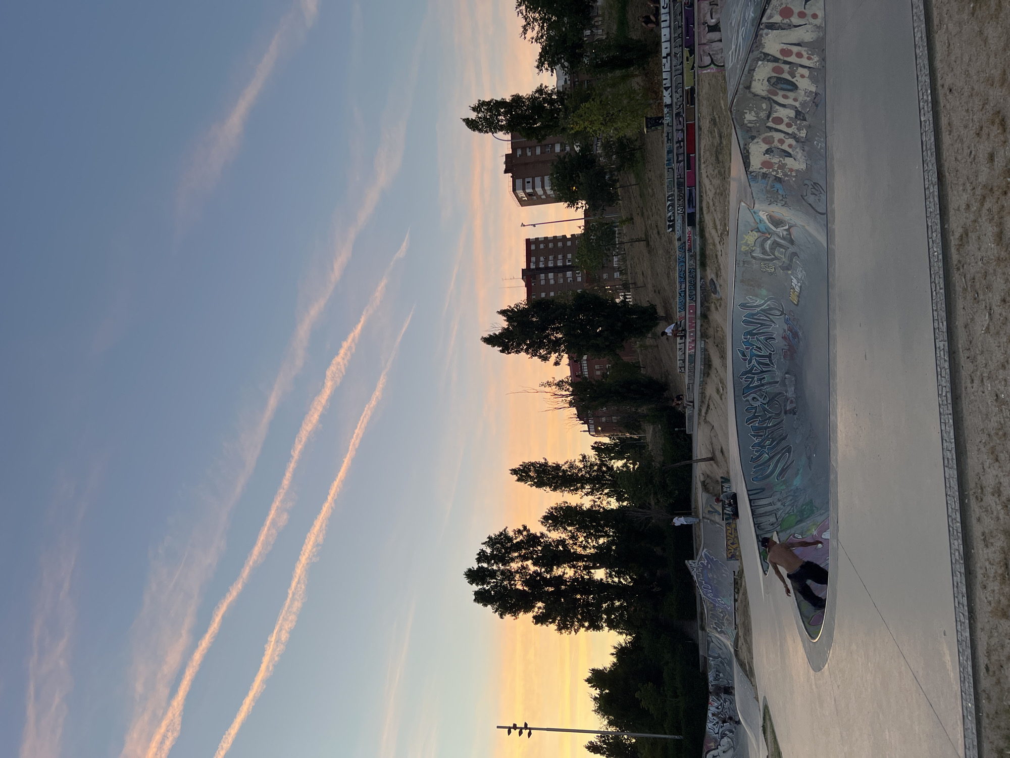 The image depicts a sunset over a skatepark in Madrid, Spain. The sky is a gradient of pale blue near the top, transitioning to a warm orange and yellow near the horizon, with faint, pinkish contrails streaking across it. In the foreground, a concrete skatepark features various bowls and ramps, many adorned with graffiti. A shirtless person is captured in mid-action, seemingly skating within a bowl. Several other figures are scattered throughout the park, some sitting on ledges, others appearing to be spectators or participants. Tall trees with dark foliage are silhouetted against the sky, and in the background, apartment buildings rise above the trees, indicating an urban setting. The overall scene suggests a late afternoon or early evening gathering at a public recreational space.