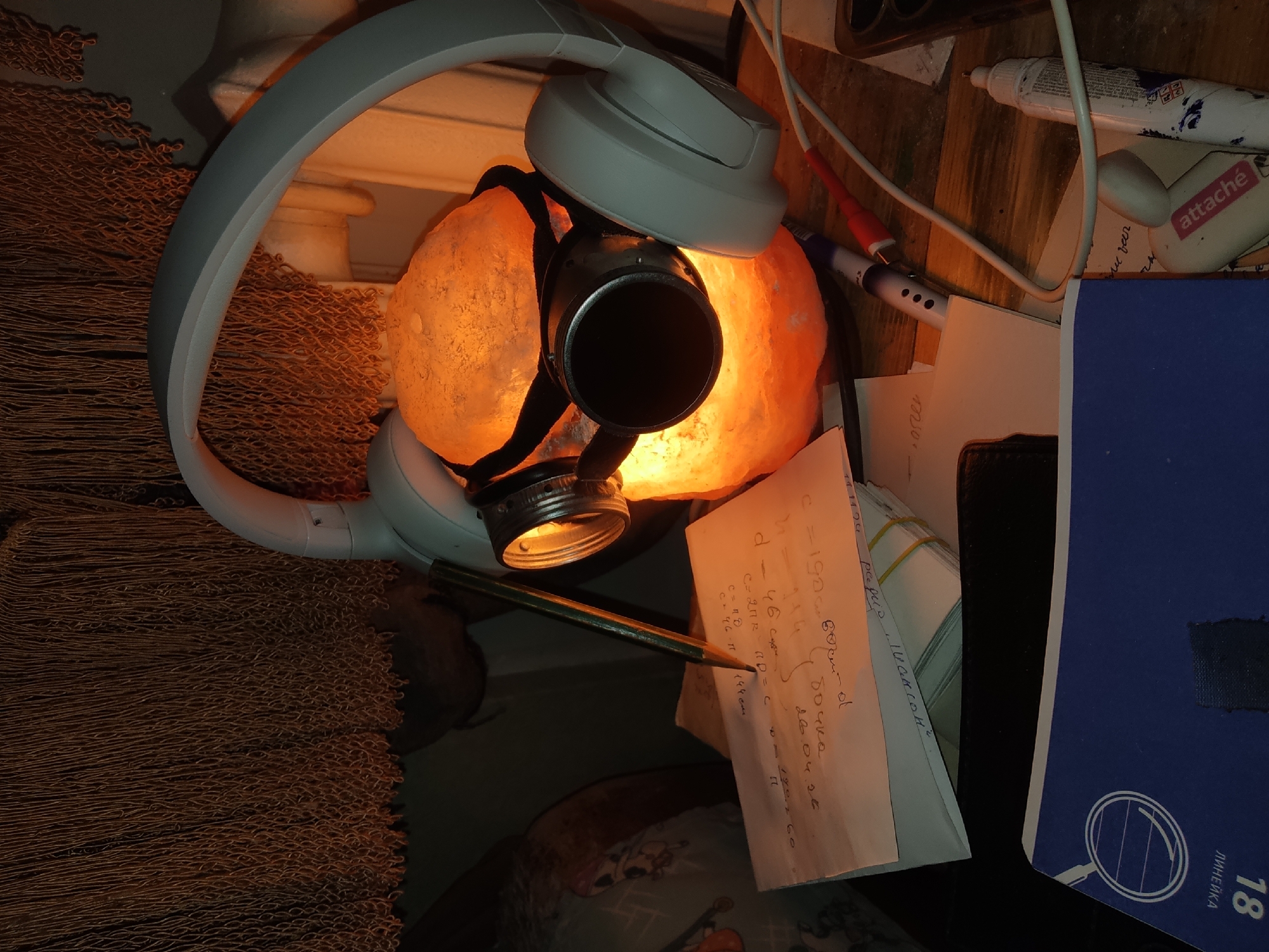 The image shows a close-up, top-down view of a cluttered desk or table. Dominating the scene is a salt lamp emitting a warm orange glow, with a black cylindrical object (possibly a camera lens) attached to it with a black strap. A pair of white headphones is draped over the lamp.
Scattered around the lamp are various items, including a piece of paper with handwritten notes and calculations, a pencil resting on the paper, a tube of adhesive (marked "attache"), and a blue notebook or folder labeled "ЛИHEЙKA" (ruler in Russian) and the number "18". Some cables and other unidentified objects are also visible. A woven brown mat/rug is located on the left. The environment is a private residence or study, Unknown, Unknown. The scene suggests a workspace or study area.