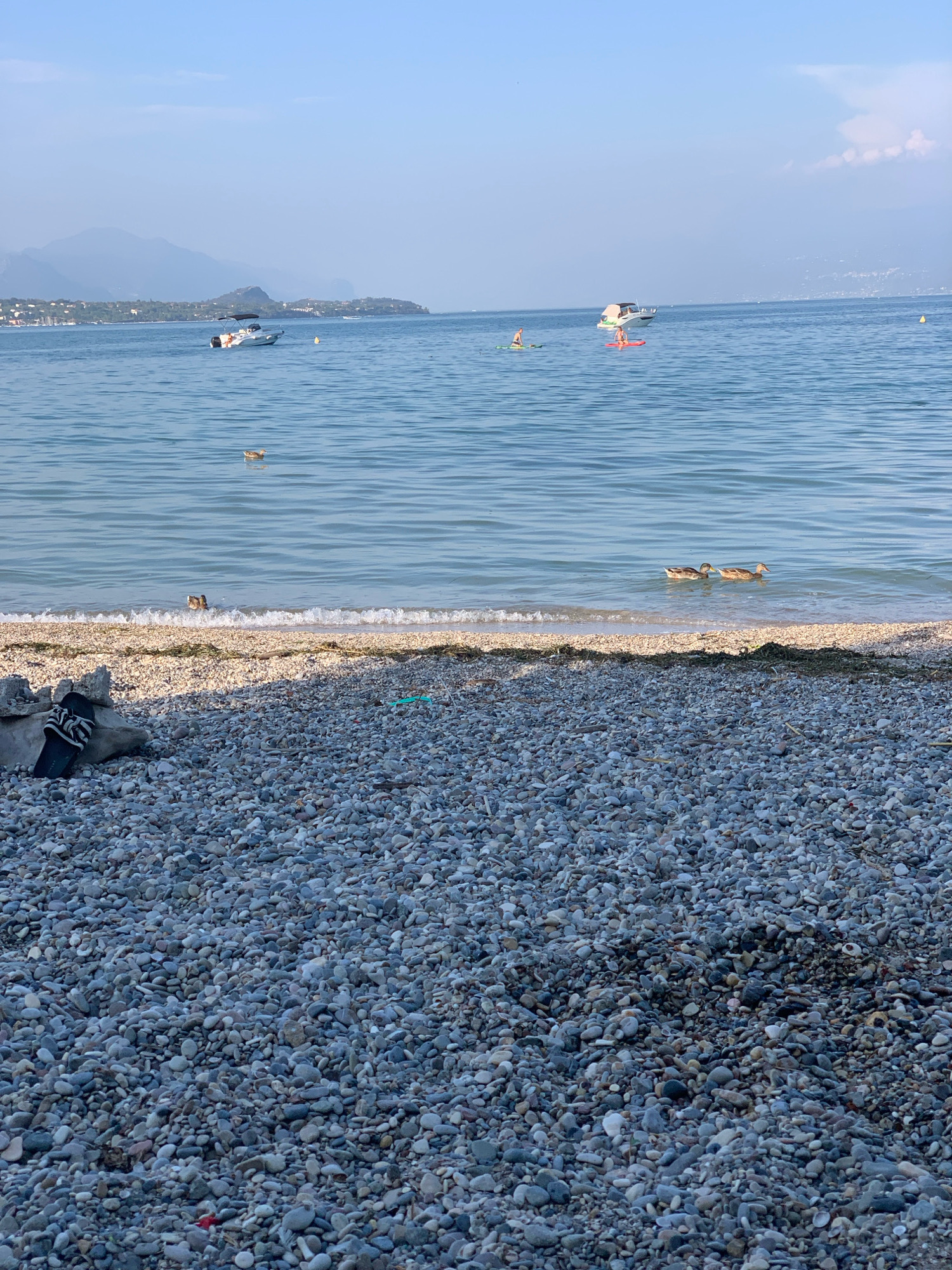 The image depicts a serene lakefront scene on a pebble beach in Padenghe sul Garda, Italy. The foreground is dominated by a vast expanse of grey and white pebbles, with a single black and white striped sandal resting on a larger grey rock to the left. In the middle ground, the calm, blue water of Lake Garda laps gently at the shore, creating a thin line of white foam. A few ducks are visible in the water, swimming close to the shore. Further out on the lake, a white motorboat with a canopy is anchored, and two individuals are seen paddleboarding. In the background, a hazy mountainous landscape stretches across the horizon under a pale blue sky with scattered clouds. The overall atmosphere is peaceful and idyllic, characteristic of a summer day at the lake.