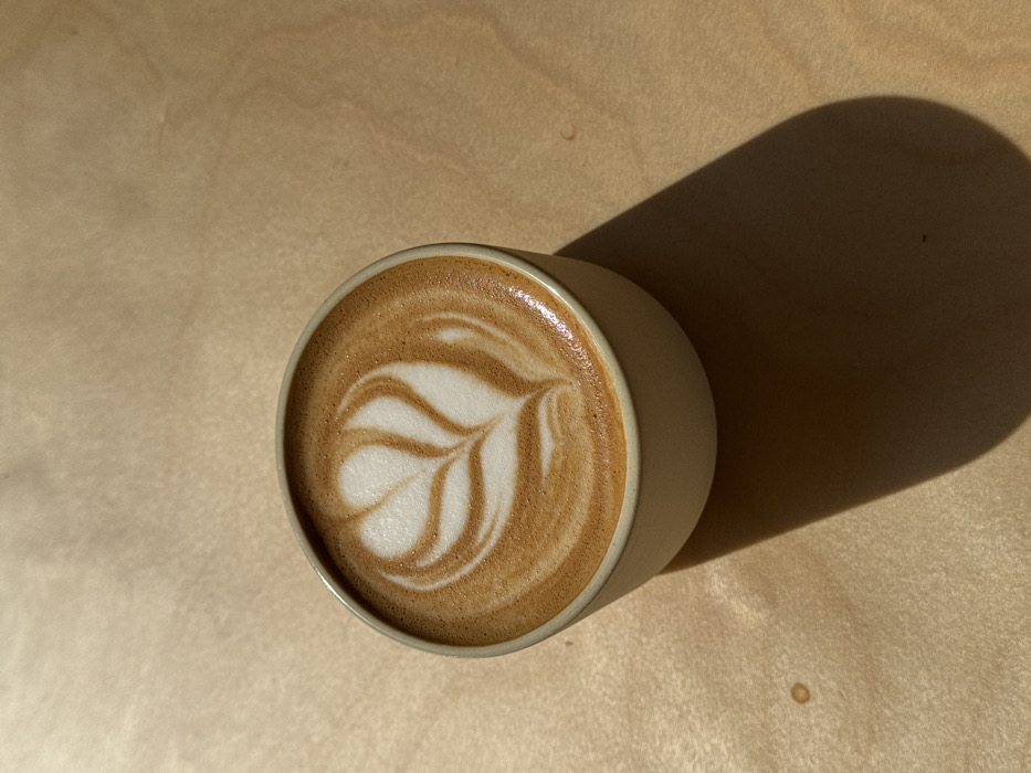 A single, light beige ceramic coffee cup is positioned slightly off-center on a smooth, light-toned wooden surface, exhibiting subtle grain. The cup contains a brown coffee beverage topped with white foamed milk, meticulously crafted into a multi-leaf rosetta latte art pattern. Strong, directional lighting from the upper left casts a distinct, elongated shadow of the cup towards the lower right of the frame. A small, faint dark circular stain is visible on the wooden surface to the lower left of the cup. The scene is located in the city of Tel-Aviv, Israel. No individuals, actions, or direct interactions are depicted.