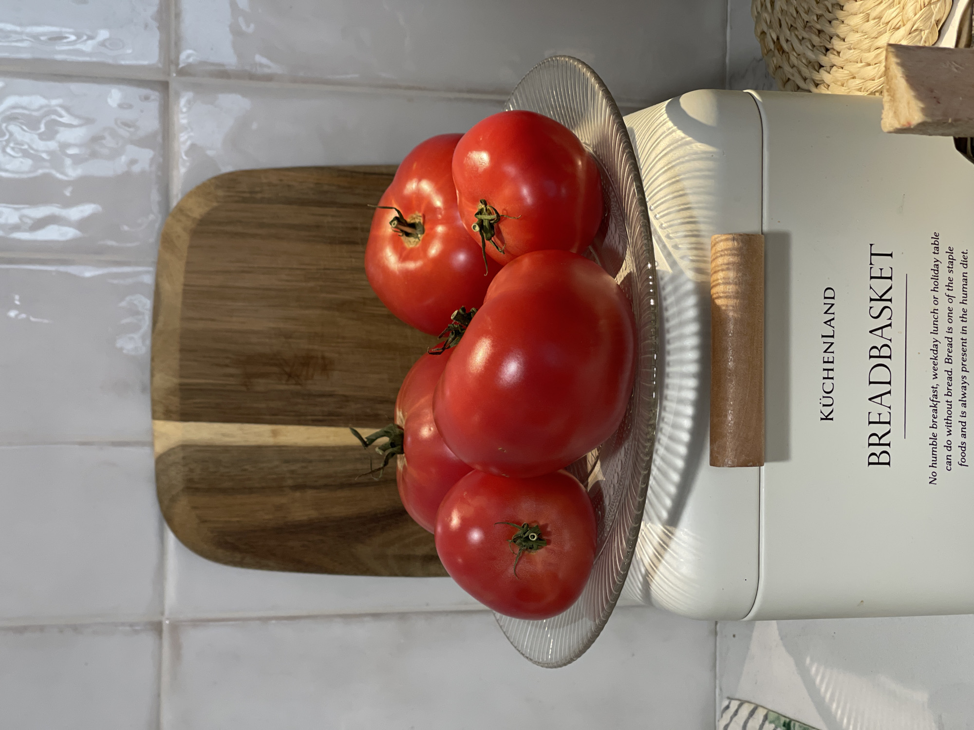 A top-down shot displays a kitchen setting with a clear glass plate containing five red tomatoes. The plate rests partly on a wooden cutting board and partly on a white bread box labeled "KÜCHENLAND BREADBASKET" with accompanying text. The scene takes place on a surface covered with white, slightly textured square tiles, possibly a countertop or table. There is also a portion of a woven basket visible on the upper right. No people are visible in the image. There is no information to indicate if this is in Izhevsk, Russia.