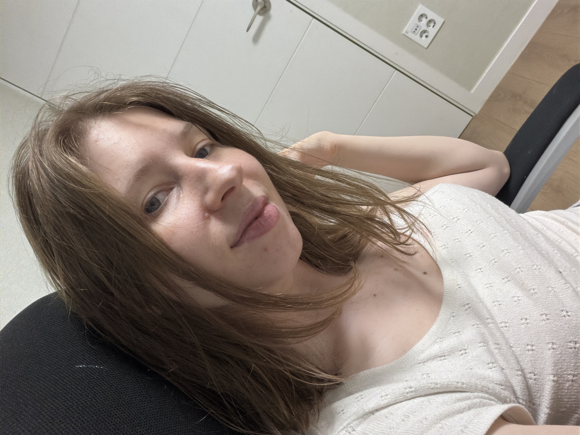 A young woman with shoulder-length brown hair and fair skin takes a selfie while sitting in a black office chair. She is wearing a white, textured t-shirt. Her eyes are open and looking at the camera, and her mouth is slightly open, with her tongue extended out. Her right arm is bent, with her hand resting on her cheek. In the background, there is a white door with a silver handle and a power outlet with three sockets. The floor is made of wood. The location is not specified as Dongducheon-si, South Korea.