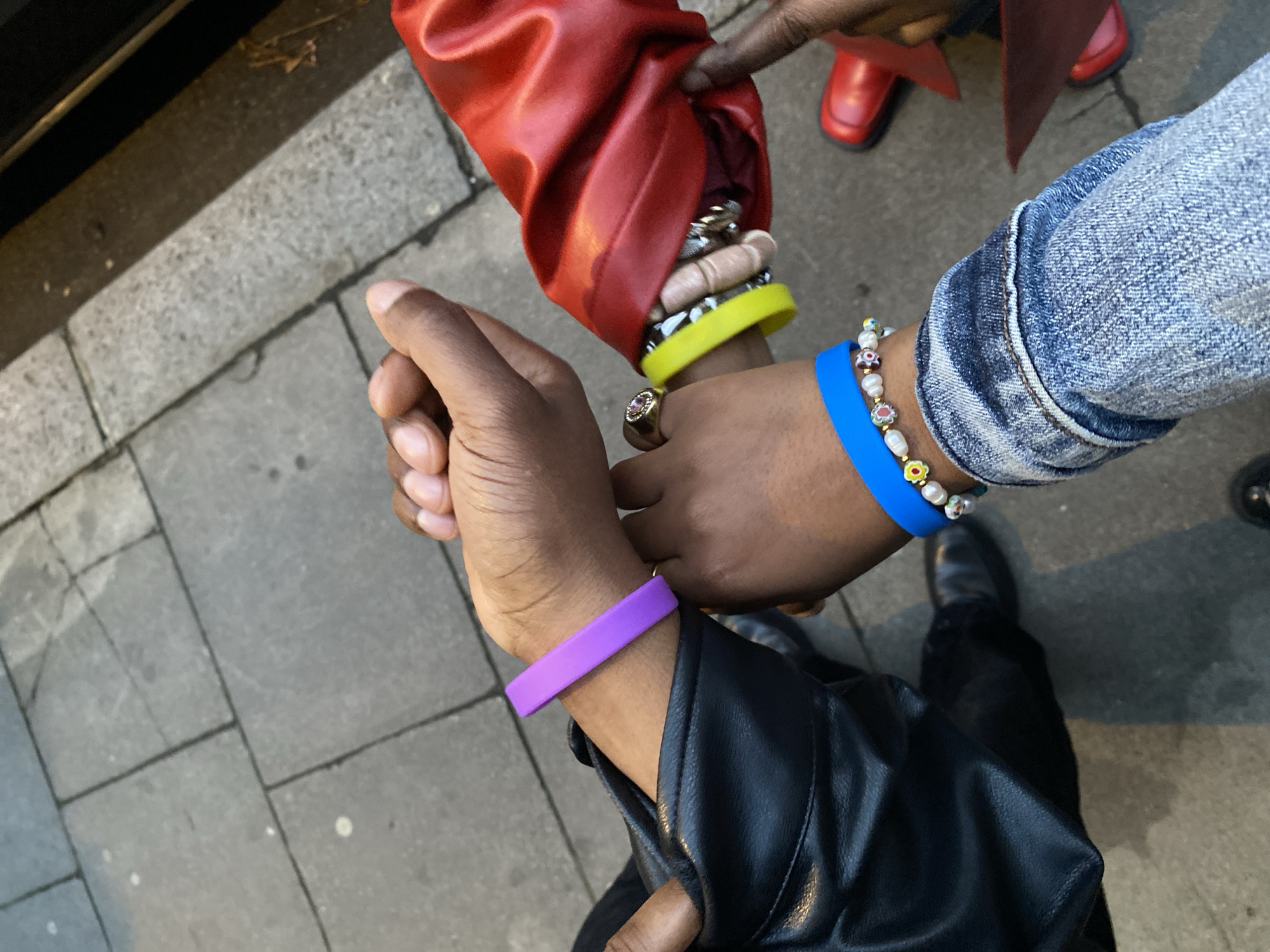 The image shows three arms, likely belonging to three different individuals, on a gray sidewalk. One arm wears a red leather jacket, a yellow wristband, and multiple rings. Another arm wears a denim jacket, a blue wristband, and a pearl and flower bead bracelet. The third arm wears a black leather jacket and a purple wristband. The ground appears to be paved with stone tiles. A pair of red shoes is slightly visible near the arm wearing the red jacket. A pair of black shoes is visible near the denim jacket arm.