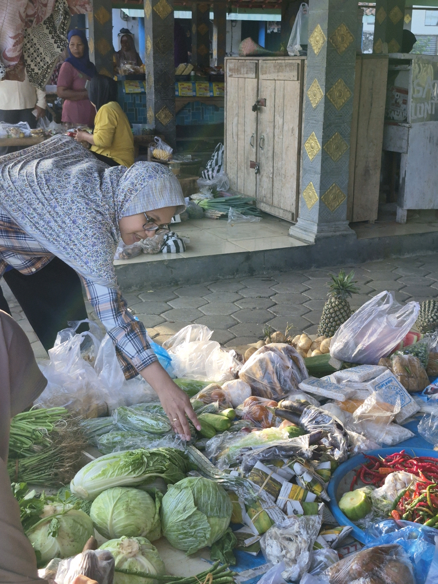 In Golok, Indonesia, a woman wearing a headscarf and glasses is in the process of shopping at an outdoor market. She is leaning forward, reaching for vegetables on a table filled with produce. The table is covered with various items, including plastic bags of vegetables, cabbages, chilies, pineapples, and other fruits and vegetables. In the background, other shoppers and market stalls are visible, with wooden structures and decorative pillars. The ground is paved with interlocking hexagonal tiles. The overall scene depicts a typical market day in Indonesia.