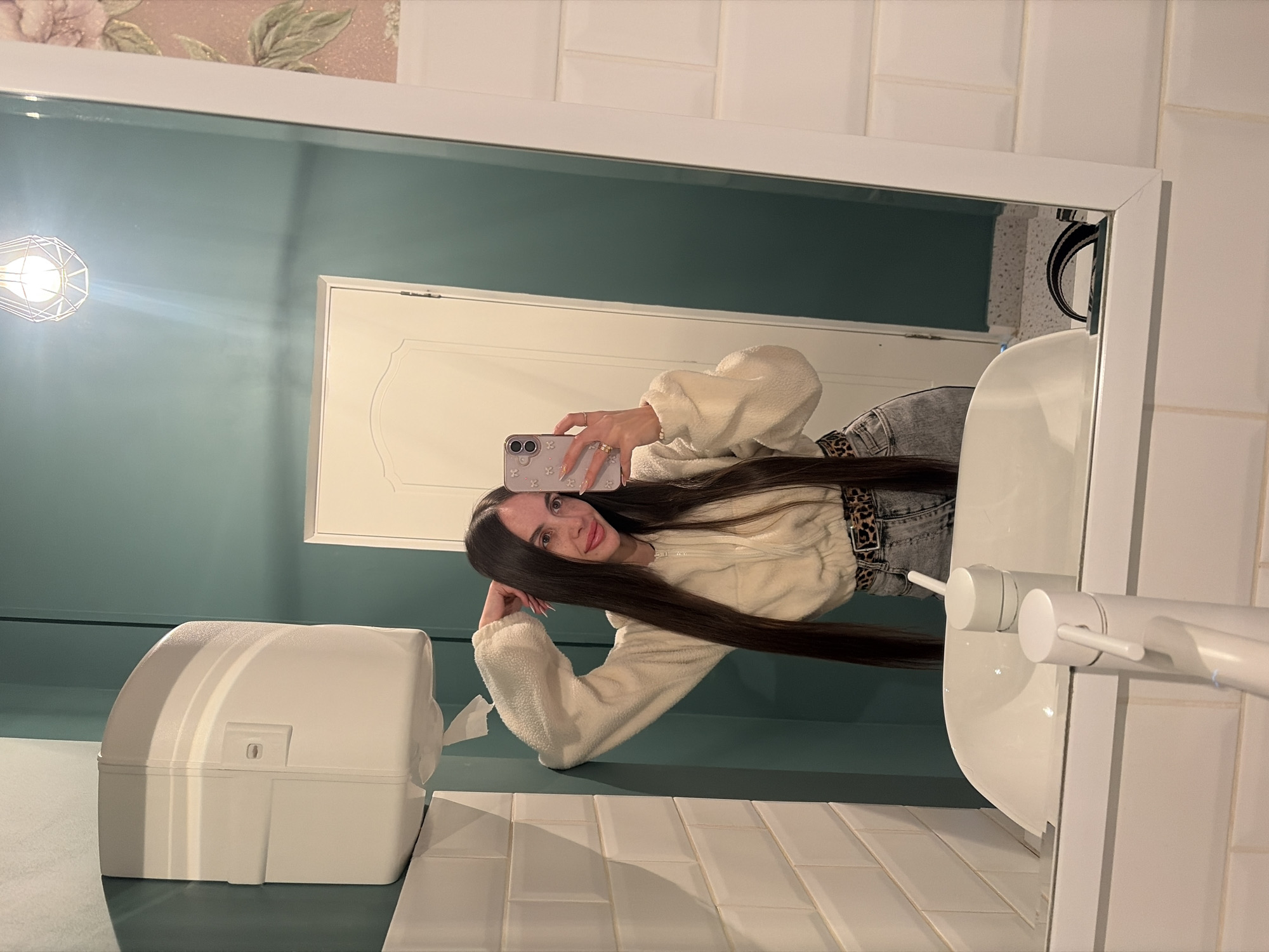 A young woman with long dark hair is taking a selfie in a bathroom mirror. She's wearing a cream-colored fuzzy jacket, and grey jeans with a leopard print belt. The bathroom has a blue-green wall, white tiles, a white sink, and a white paper towel dispenser. A light fixture is visible in the upper left of the mirror, and a door is reflected in the mirror behind her. The location is a bathroom somewhere in Saint Petersburg, Russia, as indicated by previous interaction history.