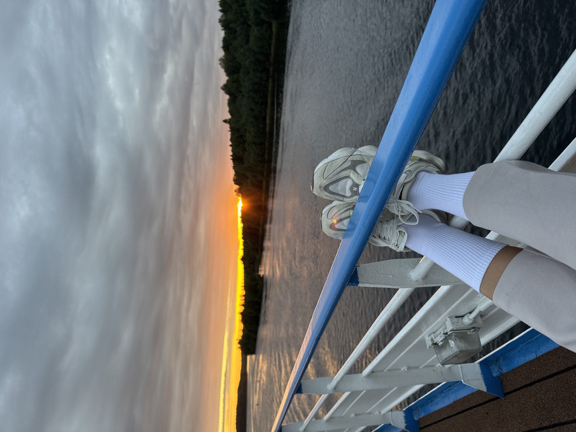 This image shows a pair of legs in white socks and sneakers resting on a blue and white railing, with a body of water and a tree-lined shore in the background. The sun is setting, casting an orange glow over the horizon and reflecting on the water. The scene appears to be on a boat or pier, given the railing and deck. The location is identified as Krasnyi Bor, Russia.