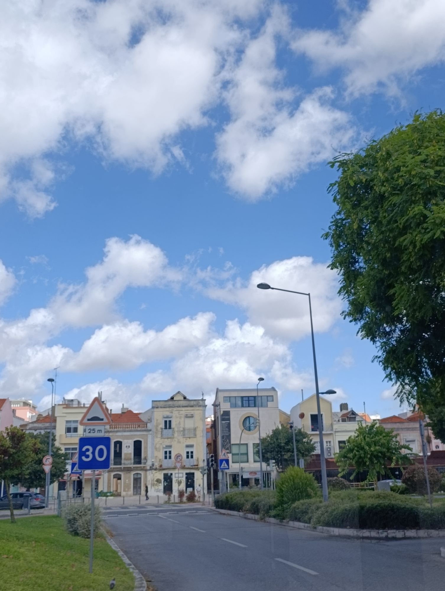 The image shows a street scene in Setúbal, Portugal, under a bright blue sky with scattered white clouds. On the left side of the road, there is a grassy area with trees and bushes. A speed limit sign indicating "30" is visible, along with a sign for a speed bump. Several buildings line the street, appearing to be a mix of residential and commercial properties with different architectural styles and colors. Some buildings have balconies and windows. A streetlamp stands on the right side of the road. A few cars are parked or driving on the street. In the background, more buildings and trees are visible. The scene appears to be an urban environment during the daytime.