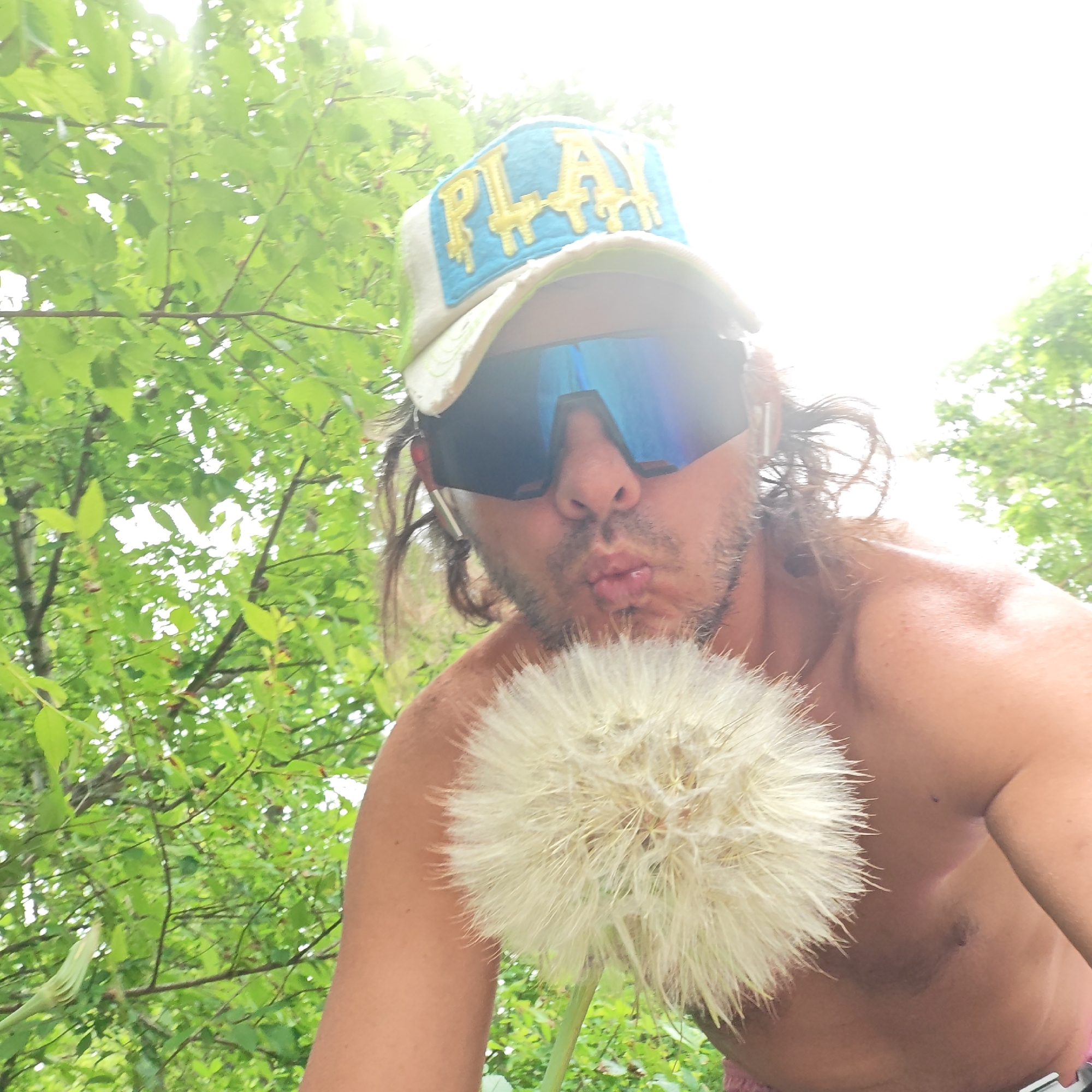A person is captured in a close-up, low-angle outdoor shot, seemingly a selfie, amidst lush green foliage. The subject is a man with long, dark hair, wearing a light-colored baseball cap with a blue and yellow "PLAY!" design, and large, reflective blue sunglasses. He is shirtless and has a beard/stubble. His lips are puckered as if to blow a kiss or to blow on something. He is holding a very large, fluffy white dandelion seed head directly in front of his face, positioned close to his mouth. The background is filled with bright green leaves and branches, indicating an outdoor, natural setting. The sky above is very bright, suggesting it is daytime and possibly sunny or overcast. The overall mood of the image appears playful and relaxed. The location context for this scene is Останинское сельское поселение, Ukraine.