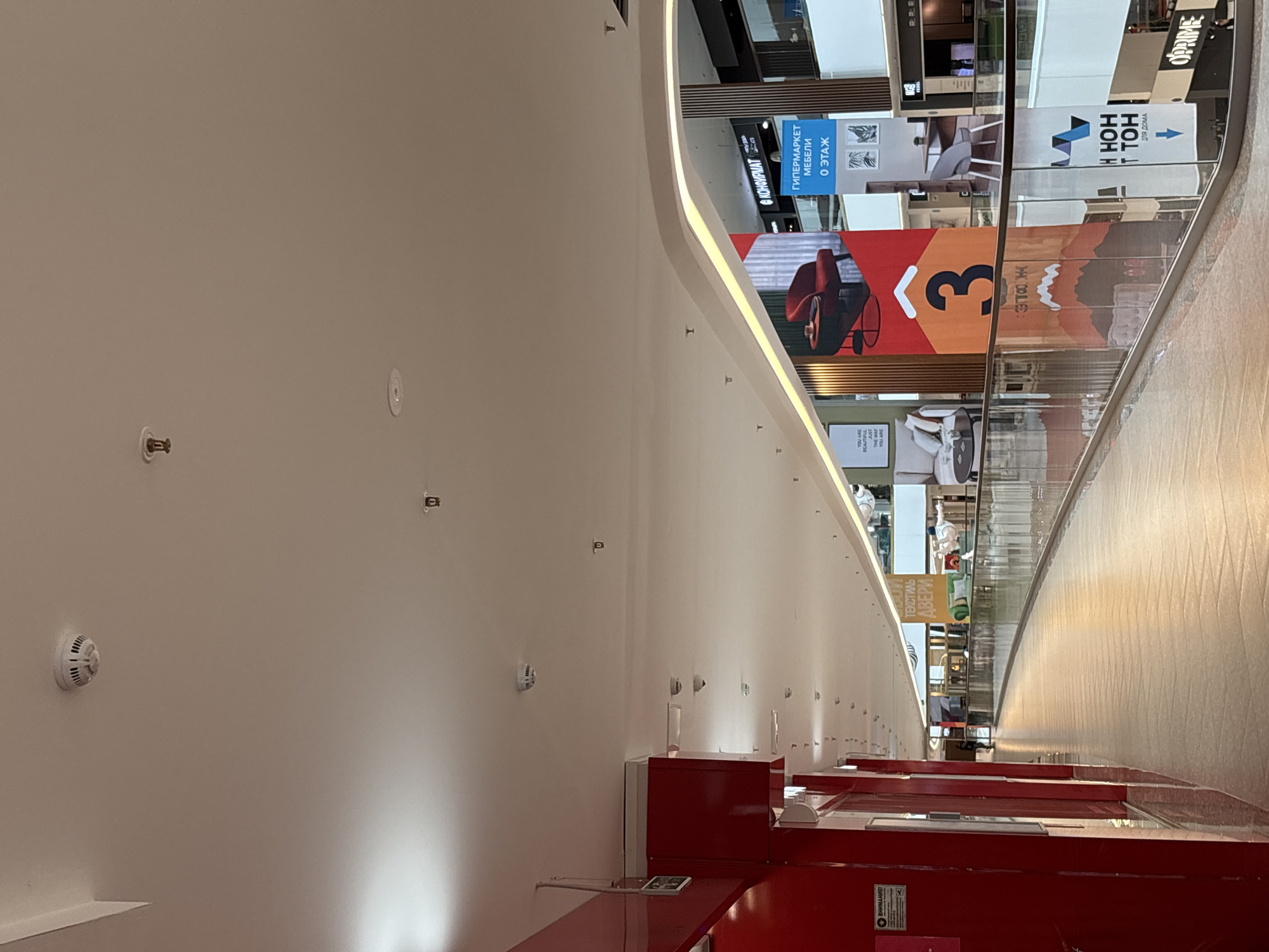 The image shows the interior of a multi-story shopping mall, likely in Saint Petersburg, Russia, as indicated by the Russian text on the signs. The perspective is upward, capturing the ceiling and upper floors. The ceiling is white with several recessed lighting fixtures and fire safety devices.
The upper floors are visible through a glass barrier, revealing advertisements and signage. One prominent sign features the number "3" in a large font and Russian text. Another sign advertises furniture ("ГИПЕРМАРКЕТ МЕБЕЛИ"). Other advertisements contain text such as "HOH TOH ДЛЯ ДОМА" and "OPRIME."
The lower part of the image shows a red structure with signs, possibly a store entrance or information desk. The overall environment suggests a modern and well-lit commercial space. No people are explicitly visible.