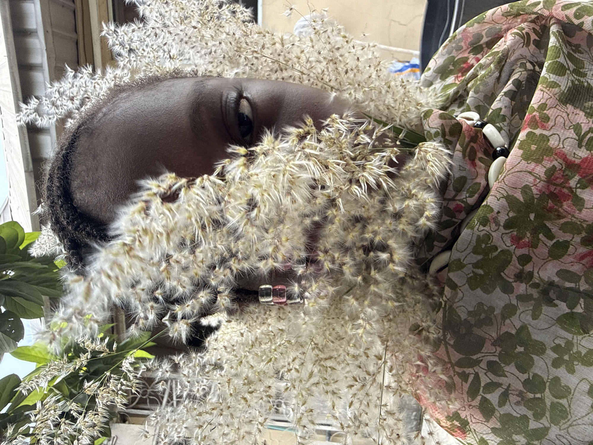 A close-up, art-directed portrait features a person with dark skin, partially obscured by an abundance of light-colored, feathery dried plant material, possibly a type of ornamental grass. The image is oriented vertically but appears rotated 90 degrees clockwise.
The subject's forehead, a single eye, and a glimpse of their mouth are visible amidst the dense foliage. Their dark, curly hair is seen around the hairline. The eye is open, looking forward or slightly upwards, conveying a calm or contemplative expression. Two small, square, translucent beads, one pinkish and one clear, are subtly visible near the person's mouth, possibly as an adornment.
The feathery plant material, predominantly light brown or off-white, covers much of the face and head, creating a soft, ethereal frame. In the bottom left corner, some vibrant green leaves add a contrasting touch of living greenery.
On the right side of the frame, a patterned fabric, likely a scarf or garment, is draped. It features a light background with a repeating pattern of green leaves and pink floral accents. Nestled within this fabric, a necklace or bracelet with alternating white and black beads, including what appear to be cowrie shells, is partially visible.
The background is largely out of focus, with hints of light-colored structures on the left and indistinct shapes elsewhere, suggesting an outdoor or brightly lit indoor setting. The natural light indicates it is daytime. The scene evokes a connection with nature and cultural adornment, placed in the context of Ado, Nigeria. No text is visible in the image.