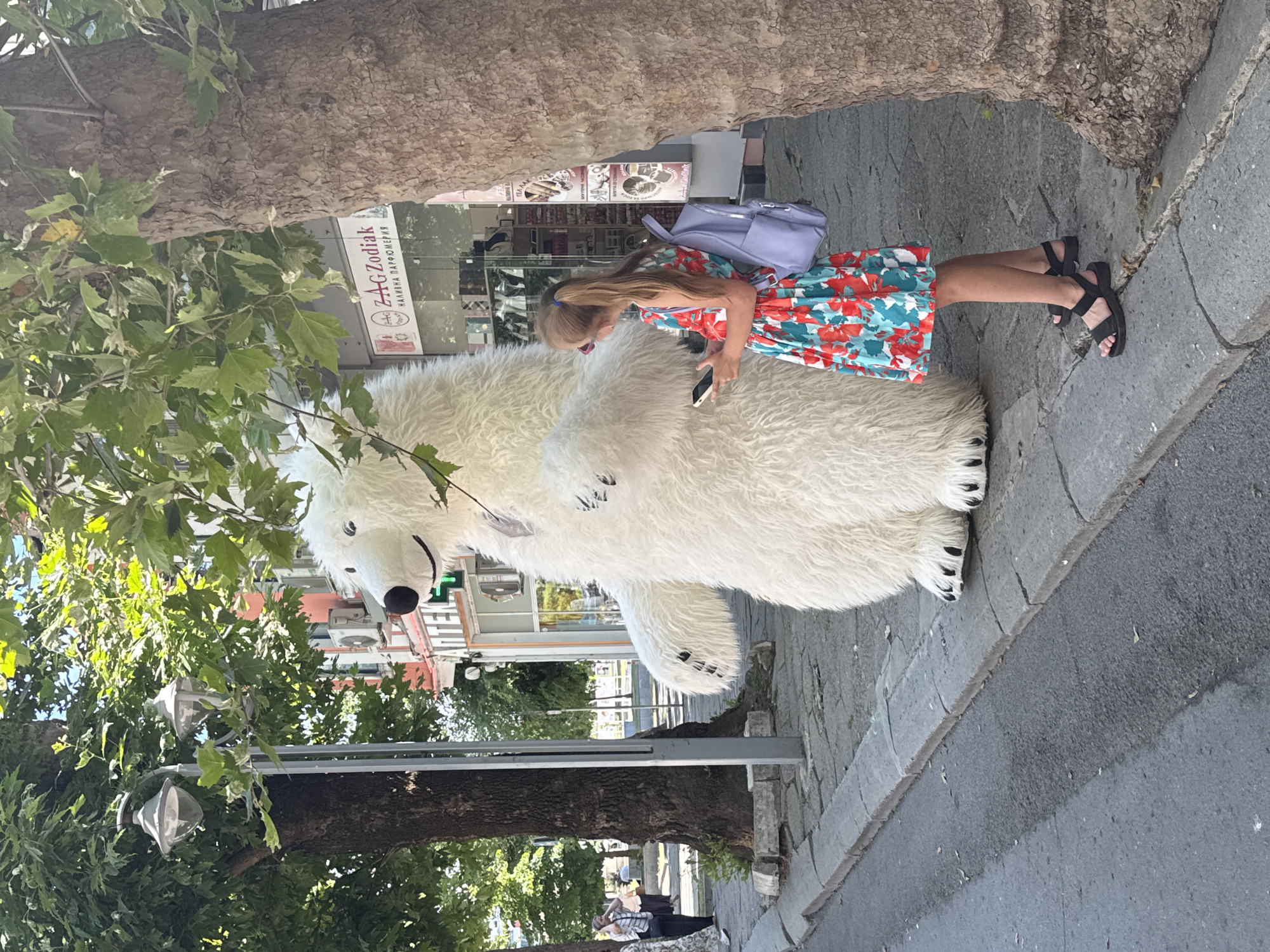 A girl in a floral dress stands next to a person in a polar bear costume on a sunny day in Burgas, Bulgaria. The girl is looking at her phone. The polar bear costume is white and fluffy, with black paw details. A purple backpack is on the girl's shoulder. The scene appears to be a street in front of shops, with trees and cobblestone pavement. Visible text on a shop sign reads "ZAG ZODIAK". The time of day is likely midday given the bright sunlight.