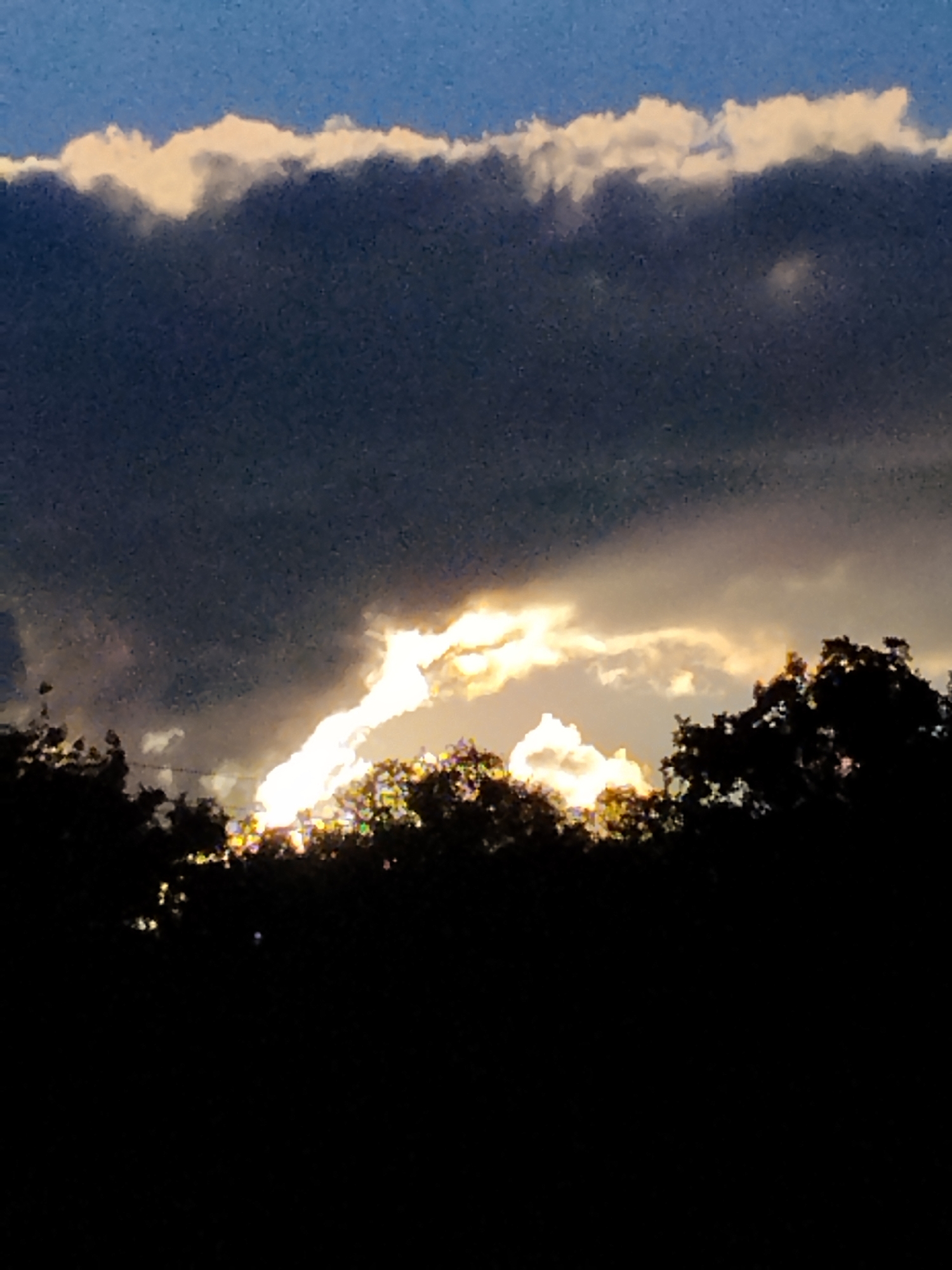 An image captures a dramatic sky over a silhouetted treeline. Dark, heavy clouds dominate the upper portion, with a bright band of illuminated clouds near the horizon, suggesting sunset or sunrise. A jagged, intensely bright area of light breaks through the clouds, casting an ethereal glow. The lower part of the image is a dense, dark silhouette of trees. The overall impression is one of natural beauty and dramatic atmospheric conditions. The image is set in Ustyvytsia, Ukraine.