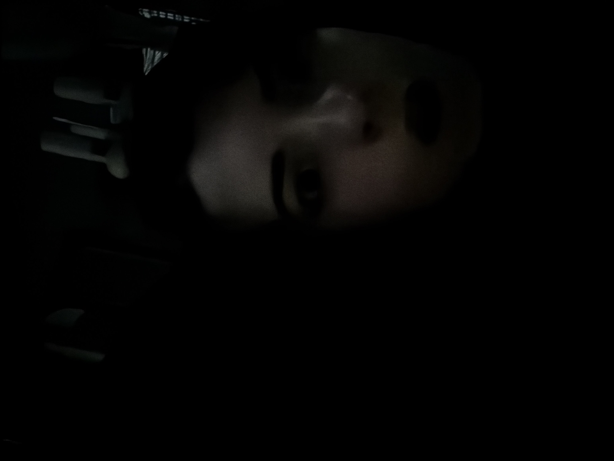 The image presents a dark, low-light portrait featuring a person, likely a woman, whose face is partially illuminated against a predominantly black background. Her head is tilted, and the left side of her face (viewer's left) is more visible, revealing an eye with a dark eyebrow, the bridge of her nose, and part of her cheek and mouth. Her hair appears dark and blends into the shadows.
To the upper left of her face, some indistinct shapes are faintly visible, suggesting fingers or a hand reaching into the frame, possibly resting on a surface or interacting with something unseen. Above her head, a faint, linear pattern hints at a structural element, possibly a ceiling vent, grill, or fan. The overall scene is enveloped in deep shadow, indicating it was captured at night or in a very dimly lit indoor environment. The sparse lighting creates a mysterious and slightly somber mood, drawing focus to the emerging facial features. No specific architectural or contextual cues for Buenos Aires are discernible directly within the image. There is no visible text.
