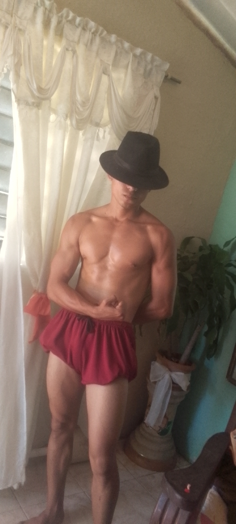 A shirtless man wearing a black fedora hat and red shorts is standing in a room. He is flexing his left bicep. Behind him are white curtains, a houseplant in a decorated pot, and a brown chair. The location is Juan Griego, Venezuela.