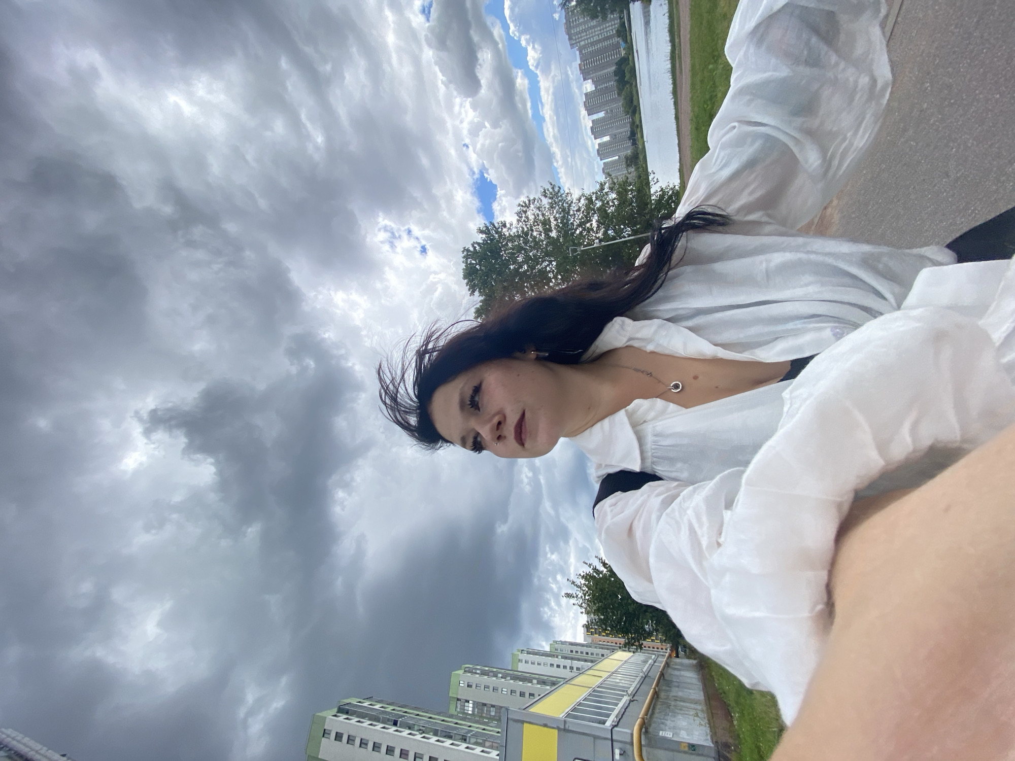In Saint Petersburg, Russia, a young woman with long dark hair stands outdoors, her face turned to the left, looking up. She is wearing a white, voluminous shirt with long sleeves, a silver necklace with a circular pendant, and a black garment underneath. Her hair is windswept. Behind her, a street is visible, lined with tall, modern apartment buildings with green accents. Further in the background, a body of water, possibly a river or canal, is present, with more apartment buildings on the far side. The sky is filled with dramatic, dark, and grey clouds, suggesting overcast or impending stormy weather, with patches of blue sky and white clouds visible towards the right. A tree is positioned between the woman and the distant buildings. The foreground shows a paved surface, likely a sidewalk or road, and a hint of the photographer's arm and shoulder. The scene captures a moment outdoors in a residential urban environment.