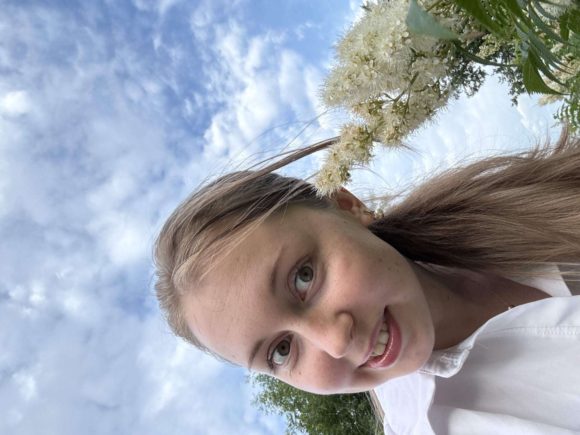 This is a selfie taken from a low angle, looking up towards the sky. The main subject is a young woman with light brown hair, smiling and looking directly at the camera. She is wearing a white collared shirt. Delicate white flowers, clustered on a thin branch, are positioned in the upper right portion of the frame, partially obscuring the sky. The background is a bright blue sky filled with fluffy white clouds, suggesting it is daytime and likely fair weather. A hint of green foliage from trees is visible at the bottom of the frame, suggesting an outdoor setting. The woman has a playful and happy expression. There is no visible text in the image.