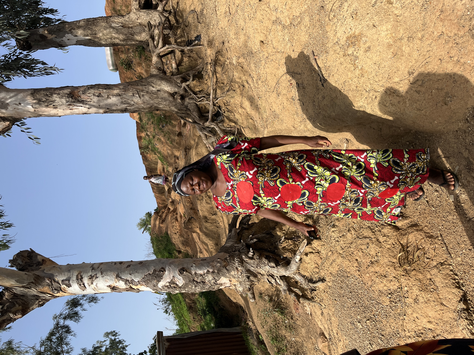 A woman in a red and yellow patterned dress stands on a dirt path, looking to her right and smiling. Behind her, a large tree with exposed roots stands on a hillside. The background shows more trees and a clear blue sky. In the distance, a small child stands on a higher elevation of the hill. The scene appears to be outdoors in a rural or semi-rural setting, potentially in Jos, Nigeria, given the typical landscape and attire.