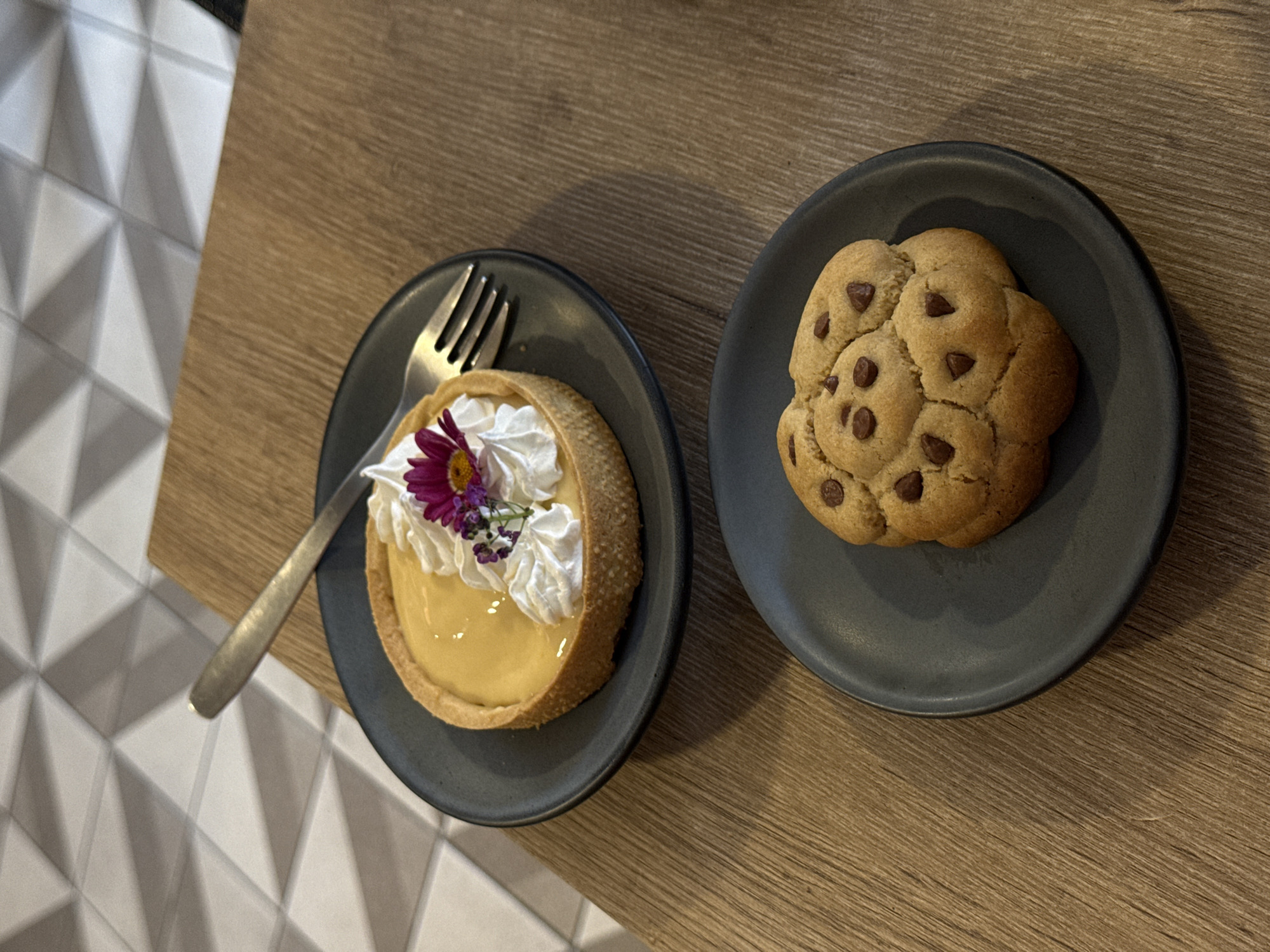 The image depicts two small, dark plates placed on a wooden-textured surface, likely a table. On the left plate, there is a small, round tart with a light yellow filling, topped with whipped cream and a small purple flower. A fork rests to the side of the tart. On the right plate, there is a single, large, round chocolate chip cookie. The background shows a section of white tile with a geometric pattern. The location is likely a cafe or restaurant in San Andrés Cholula, Mexico, based on the style of desserts and decor.