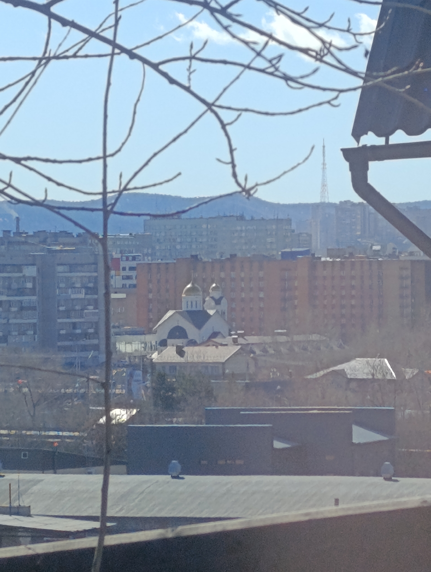 The image depicts a cityscape in Krasnoyarsk, Russia, on a clear day. In the middle ground, a white church with golden domes stands out amidst a mix of low-rise buildings and Soviet-era apartment blocks. A communications tower is visible in the distance. Bare tree branches are in the foreground and a gray roof is also visible in the lower portion of the image.