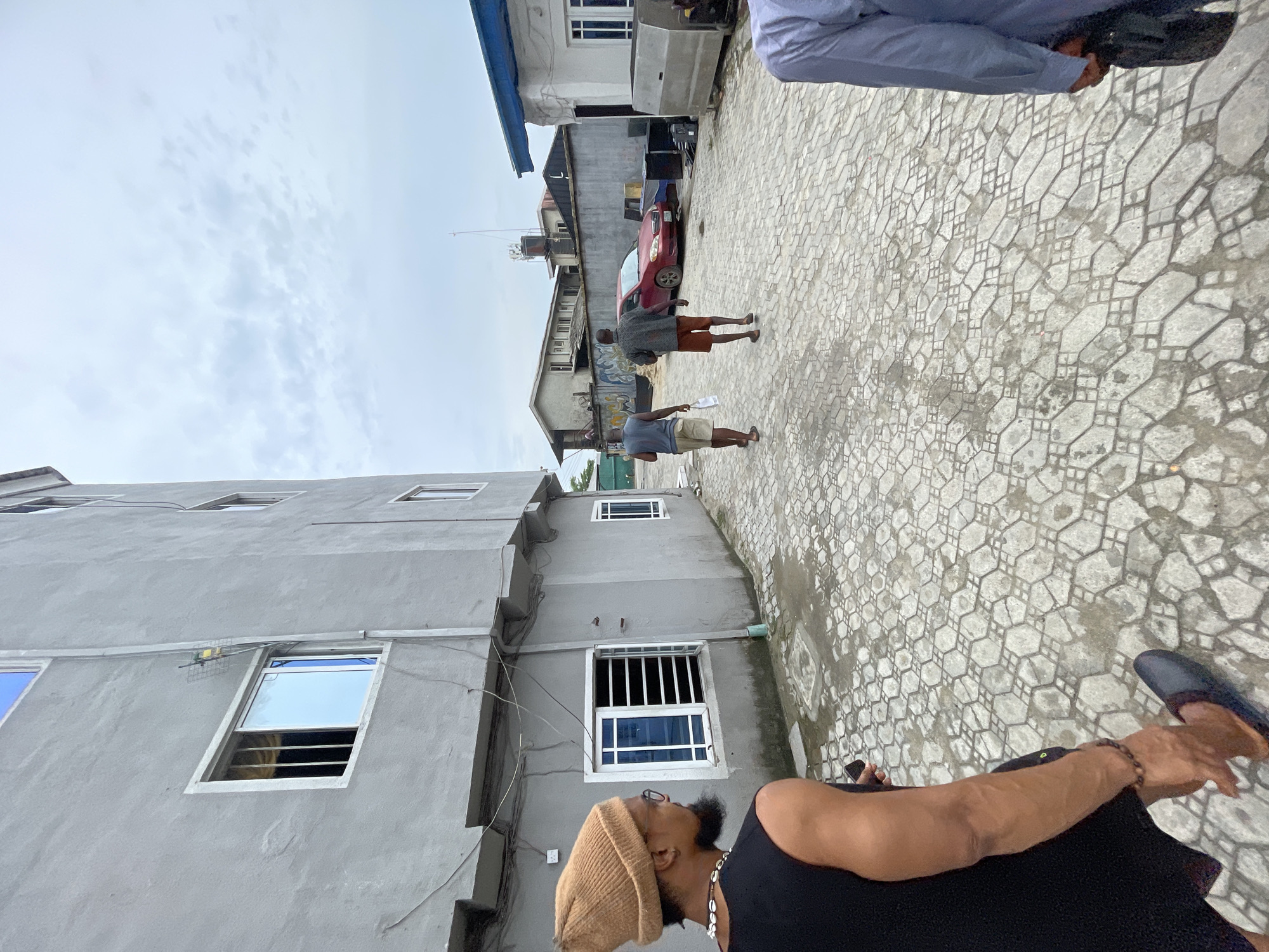 The image captures an outdoor scene in Ado, Nigeria. A multi-story, gray building with several windows, some with blue-tinted glass, stands on the left. The sky is overcast with gray clouds. Two men stand further down the concrete pavement, while a red car and other buildings are visible in the background. Another man is visible in the foreground, wearing a tan knit hat, black tank top, and beaded necklace. The pavement is composed of textured, hexagonal concrete tiles. A man in a blue shirt is on the right side of the image.