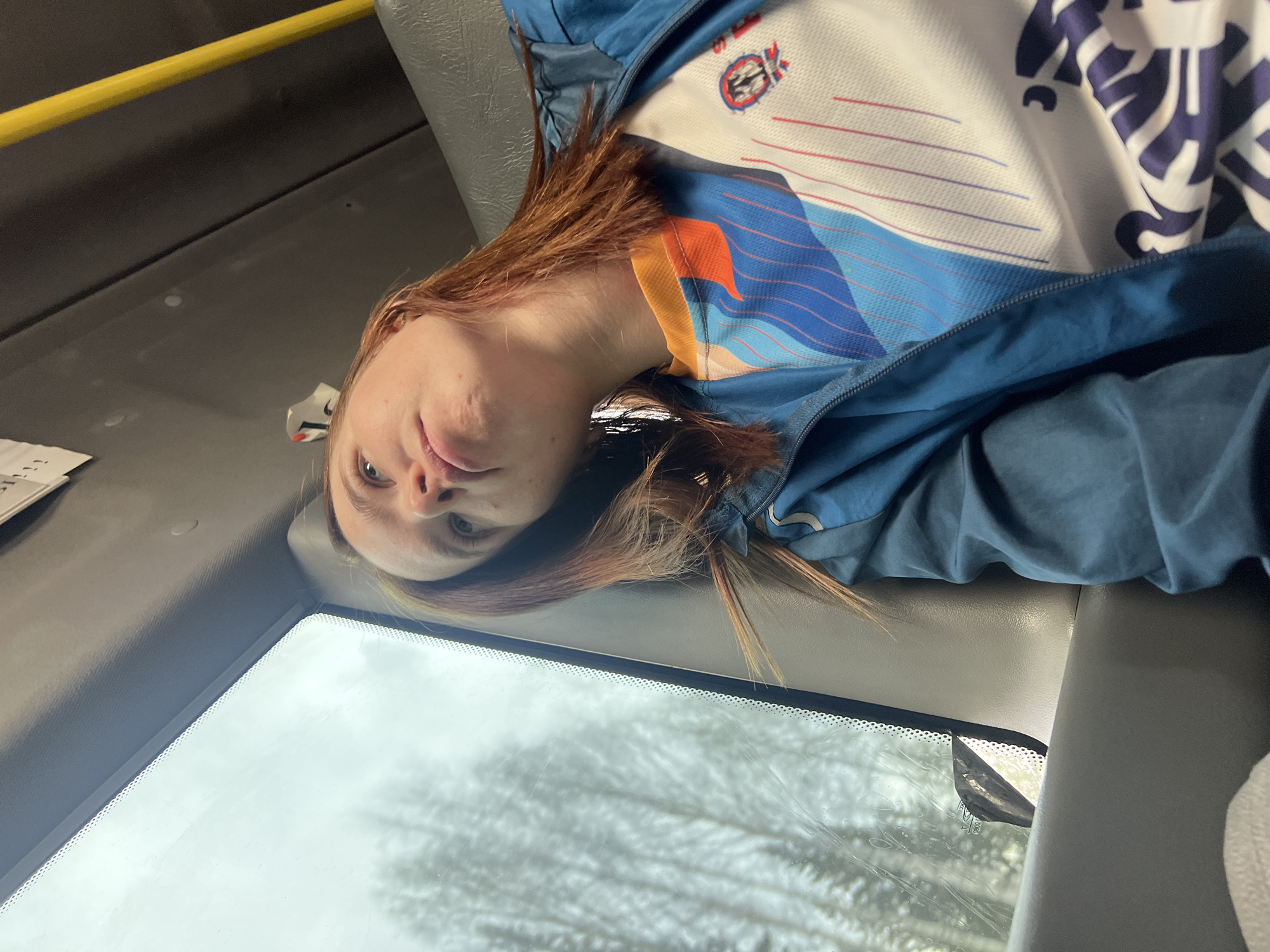 A woman with light skin and reddish-brown hair is angled diagonally in the frame, looking toward the camera. She appears to be seated inside a vehicle, possibly a bus or car, evidenced by a large front window showing a blurred outdoor scene. The exterior scene features leafless trees, suggesting a winter or late autumn setting. The interior of the vehicle includes a yellow handrail visible at the top left, and a gray interior lining. The woman is wearing a light-colored shirt with blue and white patterns, and a blue zip-up jacket. The scene suggests she is a passenger in a moving vehicle with a view of a cold, possibly overcast day. There is no information to suggest Kabanov Mys, Russia, in the scene.