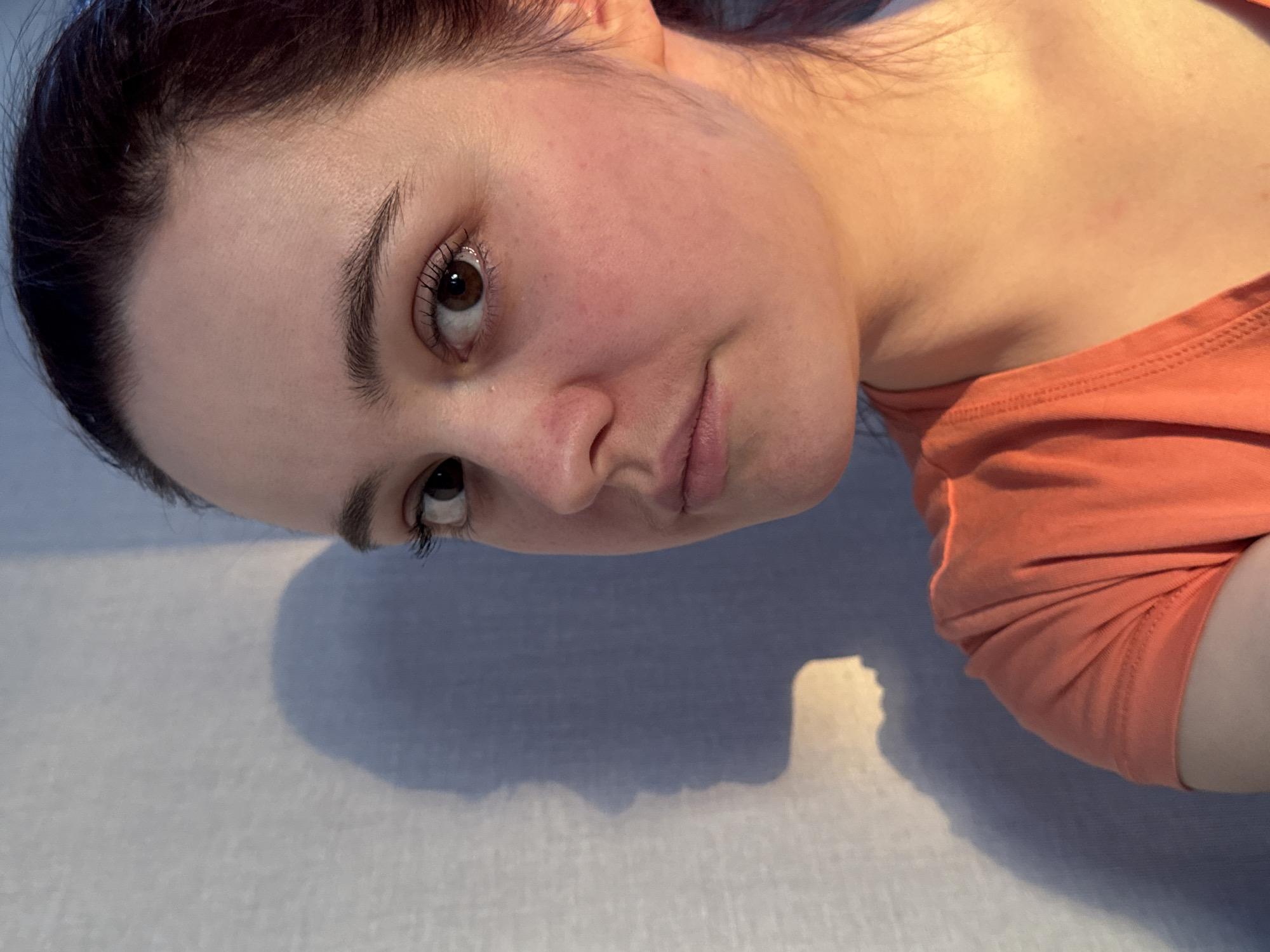 A rotated close-up portrait features a young adult, likely female, with fair skin, dark brown eyes, and dark hair pulled back from her face. She is wearing an orange short-sleeved top. The image is tilted approximately 90 degrees clockwise, with the subject's head positioned towards the lower left of the frame. She looks towards the viewer with a neutral to slightly contemplative expression, her lips hinting at a subtle smirk.
The setting appears to be indoors, against a light-colored, possibly textured fabric surface, such as bedding. Soft, warm lighting illuminates the scene, casting distinct, irregular shadows on the background, suggesting a light source from above or the side. The lighting highlights the natural texture of her skin, which shows some faint redness on the cheeks and minor marks on the neck and shoulder area. Her eyelashes are prominent, and her dark eyebrows are well-defined.
The overall impression is calm and introspective. There are no other visible objects or distinguishing features in the background. No text is visible in the image. While the provided context indicates Izhevsk, Russia, no visual cues within the image support this location.