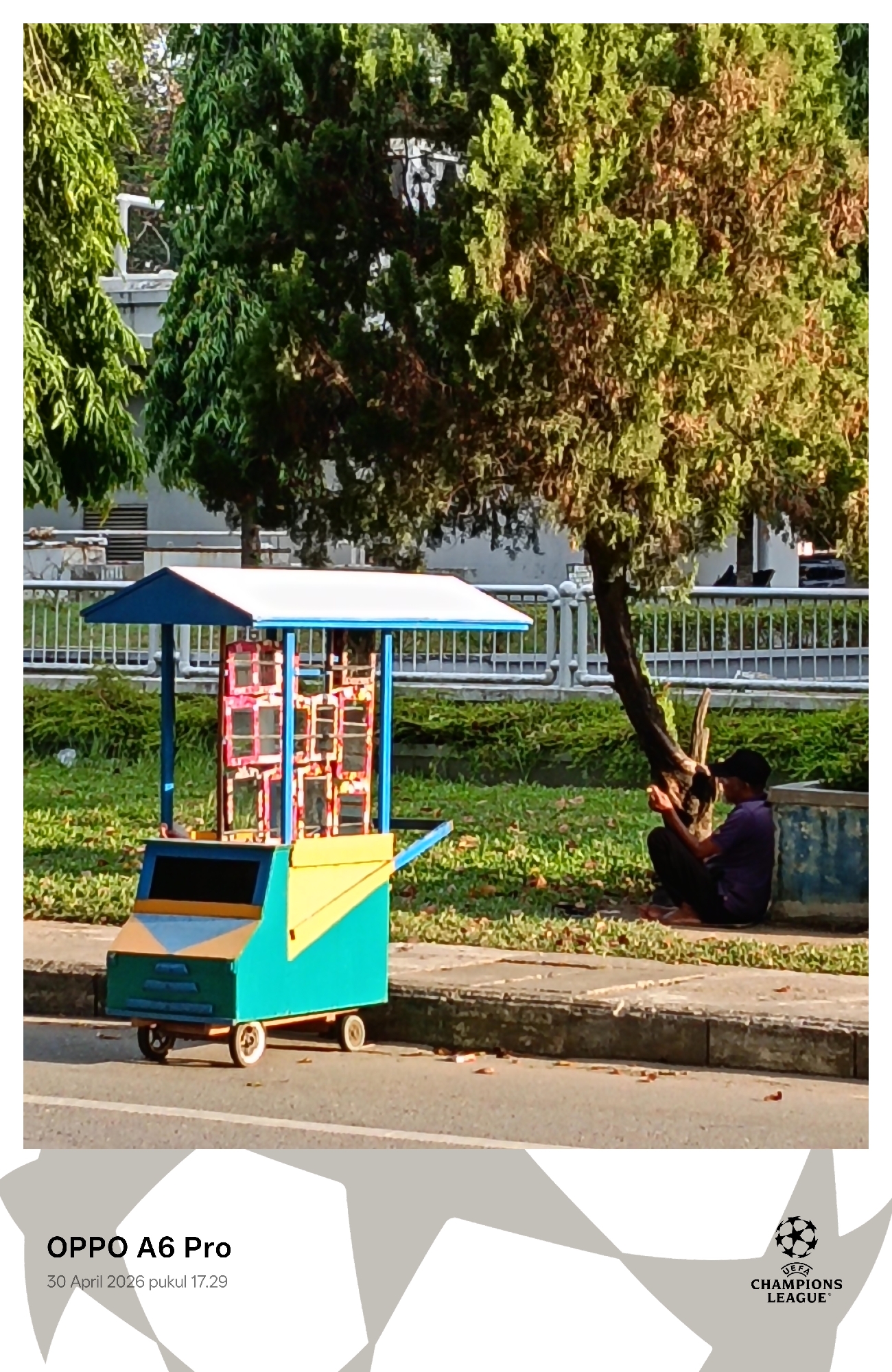 The image depicts an outdoor scene in Lhokseumawe, Indonesia, captured at 5:29 PM on April 30, 2026. In the foreground, a vibrant, multi-colored street vendor's cart is parked on the edge of a paved road. The cart has a blue roof, a teal body with yellow accents, and several small, rectangular display compartments in red and pink hues.
Behind the cart, a lush green grassy area is visible, bordered by a white metal railing in the midground. Several trees with dense green foliage, some showing hints of brown, stand prominently. To the right, a man in a dark purple shirt and a black cap is seated on the ground, leaning against the base of a tree with a distinctive, gnarled trunk. He appears to be focused on something held in his hands, possibly a mobile device.
The scene is brightly lit by the late afternoon sun, creating clear shadows and highlighting the vivid colors of the cart and the natural greenery. The overall setting suggests a public space, possibly a park or roadside, with an urban building partially visible in the distant background. The atmosphere is calm, with the man seemingly at rest.
Visible text watermarks at the bottom of the image include "OPPO A6 Pro" indicating the camera used, the date and time "30 April 2026 pukul 17.29," and the "UEFA CHAMPIONS LEAGUE" logo.