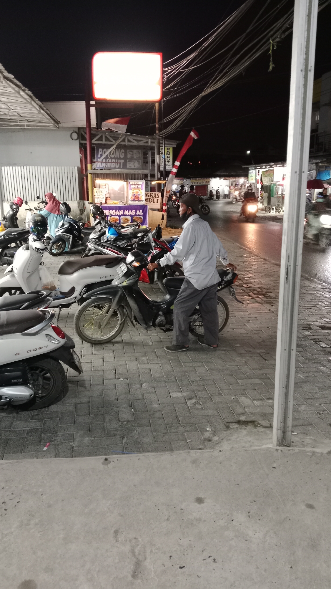 The image captures a street scene at night in Handil Bakti, Indonesia. A man in a light-colored shirt and dark pants, wearing a dark cap and a mask, is standing next to a motorcycle, leaning on its handlebars. The motorcycle is parked on a paved walkway made of interlocking bricks. Several other motorcycles are parked nearby, some partially obscured. Behind the motorcycles, a food stall with a banner advertising "POTONG RAMBUT" (haircut) and "WARUNG MAKAN" (food stall) is visible. An illuminated sign with red trim is mounted above the stall. The street in the background is wet, reflecting the lights of passing vehicles. A few people are seen on motorcycles in the background, and a red and white flag is displayed on a pole near the sign. The overall environment appears to be a commercial area with street vendors.
