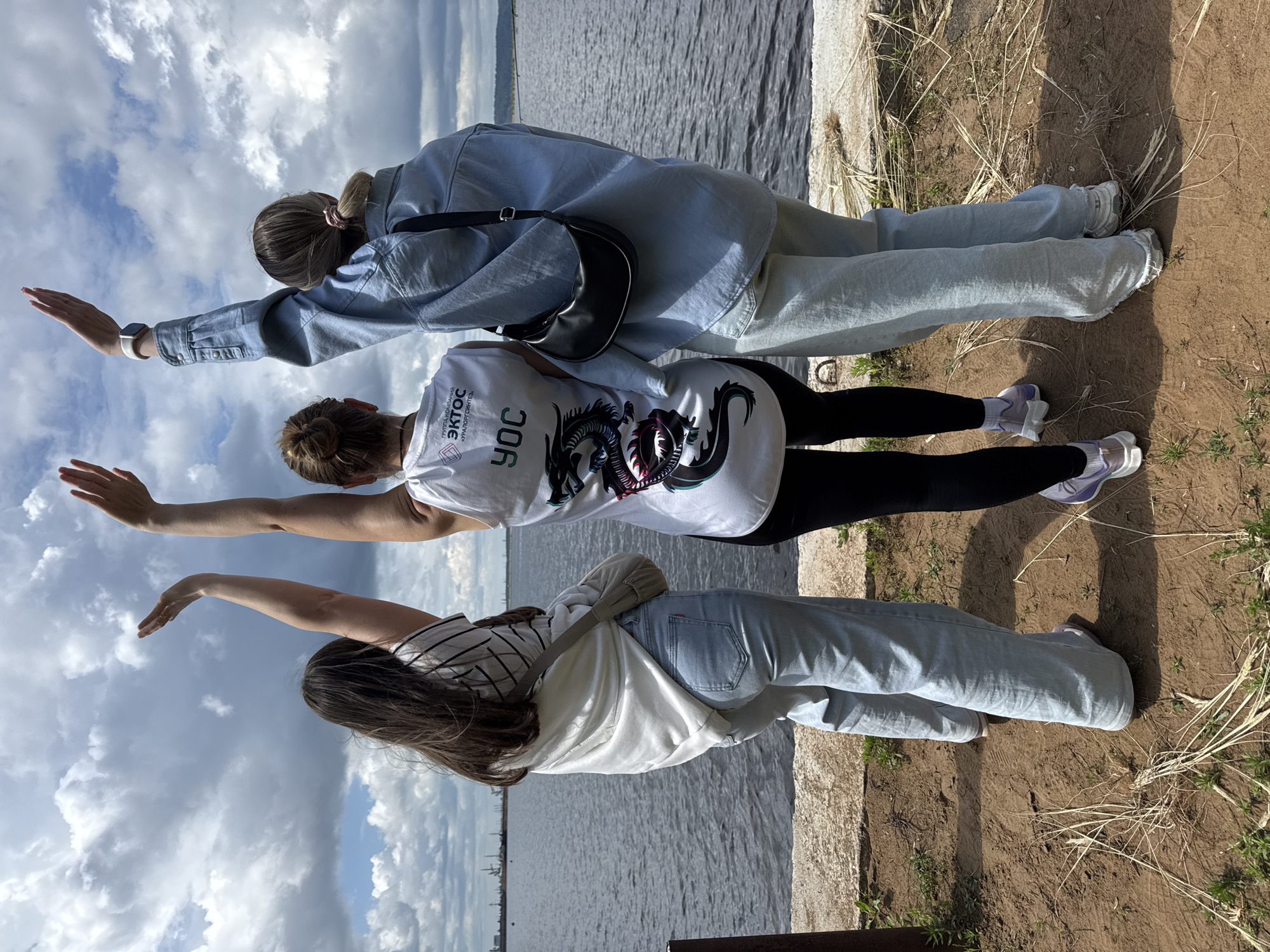 Three young women stand with their backs to the camera, facing a body of water under a cloudy sky. They are positioned on a sandy, slightly overgrown embankment, with some dry grass and green shoots visible. The woman in the center wears a white tank top with a dragon graphic and text that appears to be in Russian. The woman to her left wears a striped long-sleeved shirt and light-wash jeans. The woman to her right wears a light blue denim jacket over a light-colored shirt and light-wash jeans. All three women have their right arms raised in a celebratory gesture. In the background, the water stretches to the horizon, where industrial structures and a faint tree line are visible. This scene is likely located along the Kama River embankment in Chaikovsky, Russia, a city known for its hydroelectric power station and proximity to the river.