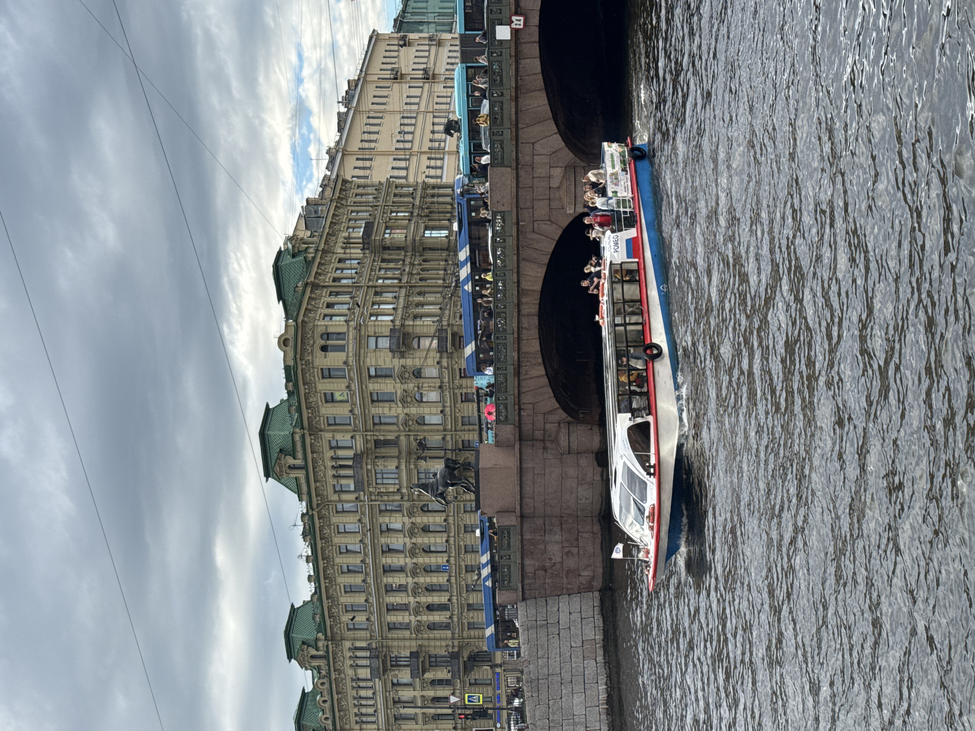 The image captures a boat tour on a canal in Saint Petersburg, Russia. The boat, adorned with red and blue stripes, is filled with passengers enjoying the sightseeing. It is moving past a canal bank and a large, ornate building with a green roof, typical of St. Petersburg's architecture. The building has numerous windows and intricate detailing, suggesting historical significance. Underneath a bridge, the water reflects the overcast sky, indicating a cloudy day. Power lines are visible stretching across the sky, adding to the urban landscape. A statue of a man on horseback is prominently displayed on a pedestal near the canal bank, adding to the historical ambiance of the scene. The overall mood is one of calm observation and sightseeing. The word "POMEG" is visible on the side of the boat.