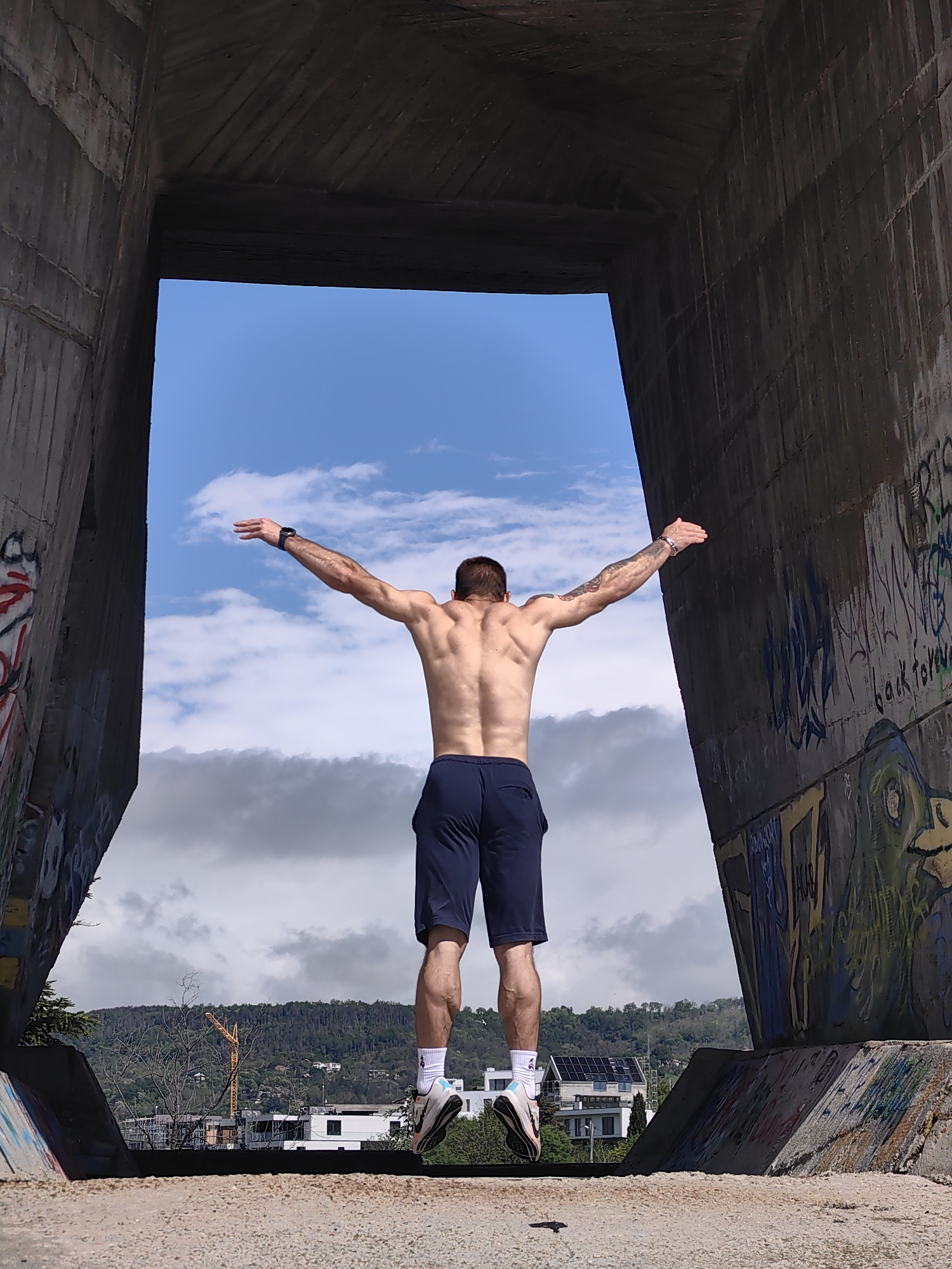 A shirtless man wearing dark shorts and white socks is jumping in the air with his arms outstretched. He is positioned within a concrete structure, possibly part of an unfinished or abandoned construction project, that frames a view of the sky and the cityscape of Varna, Bulgaria. The concrete structure features graffiti on its surfaces. In the background, there are buildings, trees, and a construction crane visible against a cloudy sky. The perspective is from a low angle, looking up at the man as he jumps.