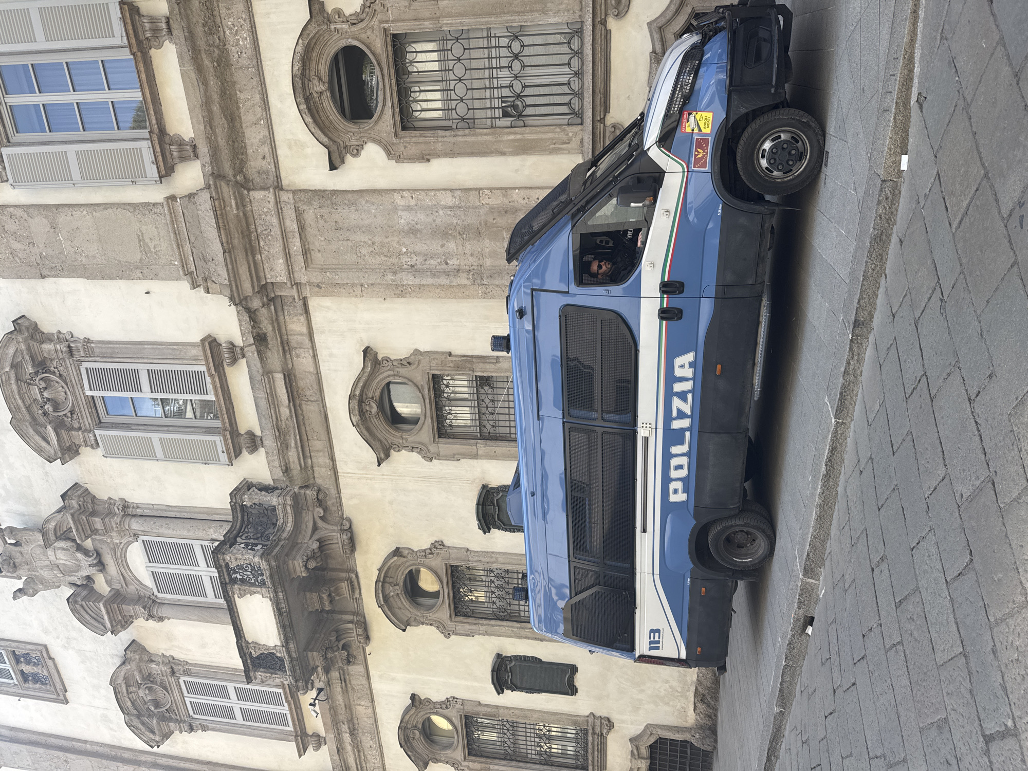 A blue and white Italian police van is parked on a paved street next to an ornate building. The van is positioned such that its side is facing the viewer, displaying the word "POLIZIA" in large white letters. The building facade features multiple windows, some with closed shutters and others with decorative metal bars. The architecture suggests a historic location, likely in the city of Milan, Italy. A person can be seen in the driver's seat of the van. The surroundings suggest an urban environment with traditional European architectural elements.