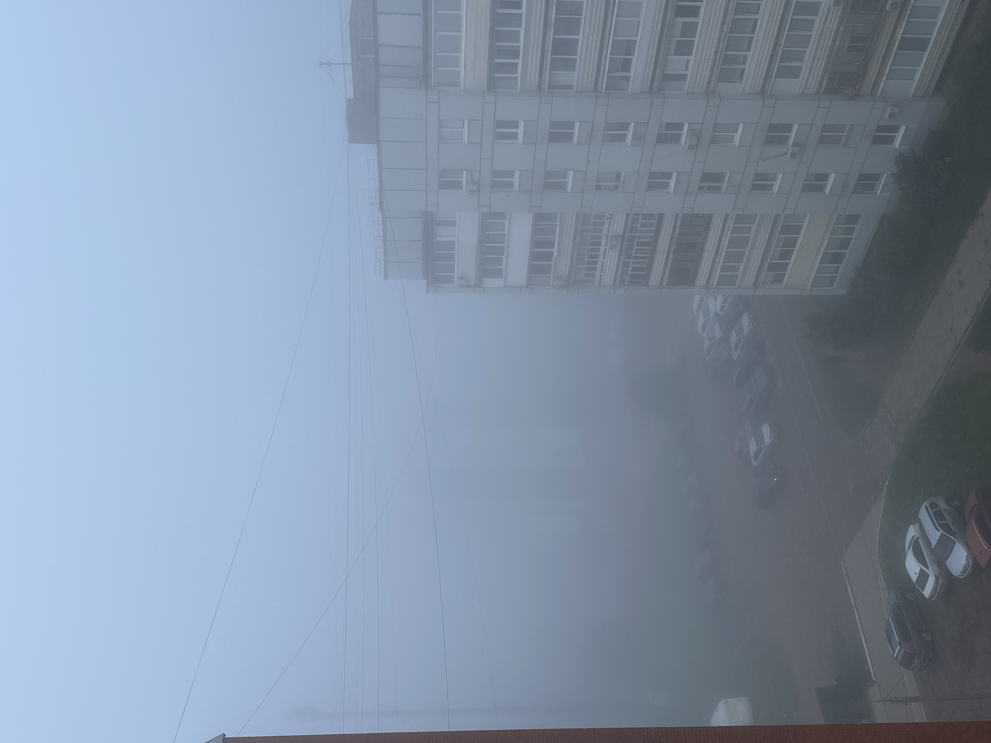 An outdoor scene is depicted from a high vantage point, looking down into a foggy urban landscape. The image is taken from above a reddish-brown brick structure, possibly a wall or roof edge, which runs along the bottom of the frame.
The dominant feature is the thick, pale blue-grey fog or mist that permeates the entire scene, significantly reducing visibility. Through the fog, a large, multi-story building occupies the left side of the frame. This building, likely an apartment block, is light-colored with numerous rectangular windows and visible architectural details, including ledges and what appear to be air conditioning units on its exterior. Its upper sections are particularly obscured by the haze.
To the right and below the building, a parking lot is visible, with several cars parked on a paved surface. Among them, white cars are prominent, along with one darker vehicle and a glimpse of a red/orange car. Adjacent to the parking area are patches of green grass and a paved pathway that winds through the misty environment.
Numerous thin, dark utility wires or cables stretch diagonally and horizontally across the upper and middle portions of the sky and towards the building, disappearing into the fog.
The weather is extremely foggy, creating a muted and somewhat indistinct atmosphere. The ambient light suggests it is daytime, though the exact time is difficult to ascertain due to the heavy mist. There are no people visible, and no specific activities are taking place beyond the static scene. No visible text is present in the image. The general setting suggests a residential or urban area, characteristic of post-Soviet architecture, which aligns with the provided location context of Chaikovsky, Russia.