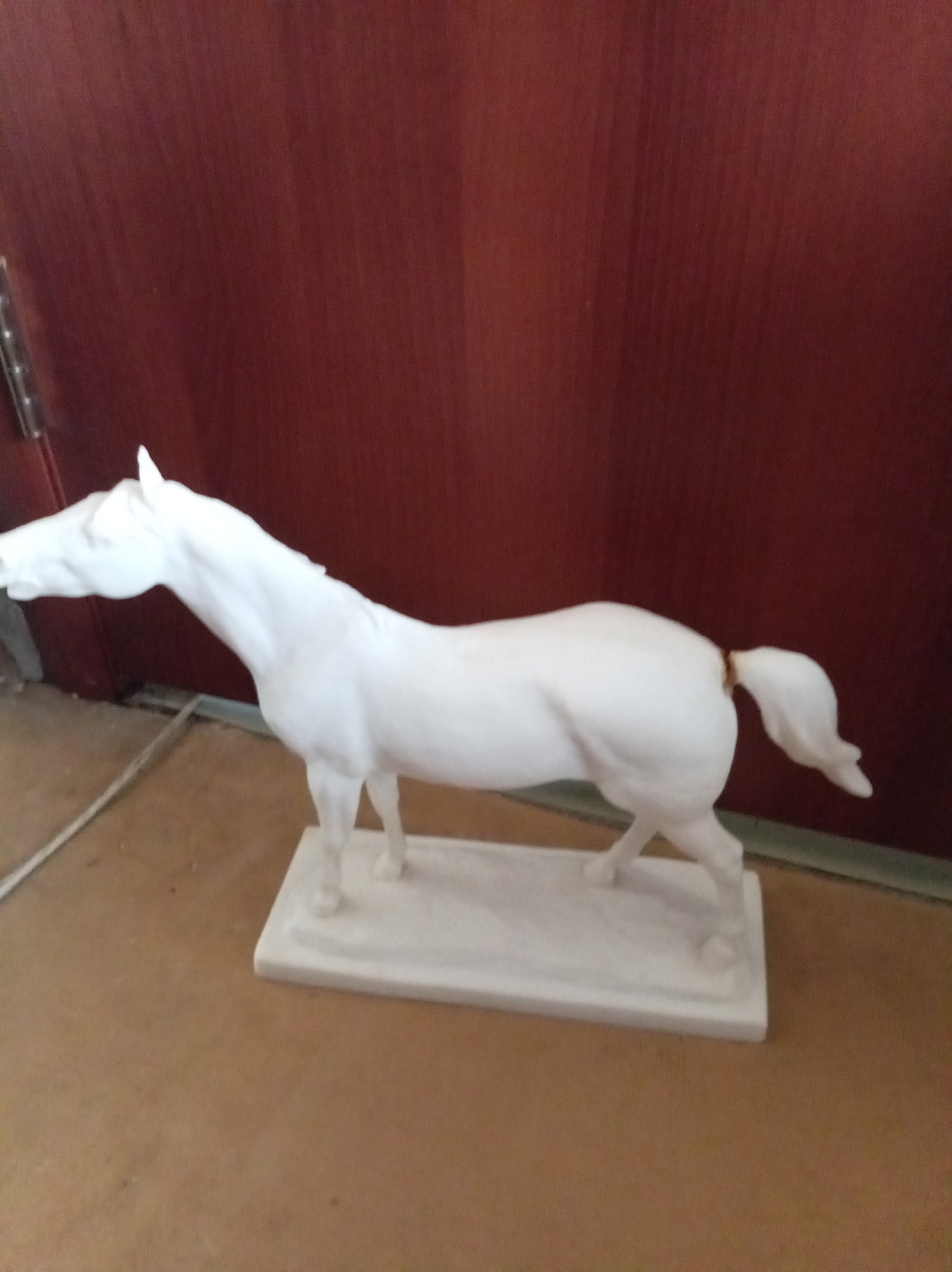 A white porcelain horse figurine stands on a rectangular base. The horse is depicted in profile, facing left, with its head lowered and its body in a natural stance. It is positioned on a flat, light brown surface, likely a floor or table. Behind the figurine, a dark red, textured surface, possibly a wall or door, serves as the backdrop. The lighting appears to be from an overhead source, casting subtle shadows. The environment suggests an indoor setting, possibly a home or a display area. The figurine's smooth, unadorned finish emphasizes its form. There are no people or other animals visible in the image.