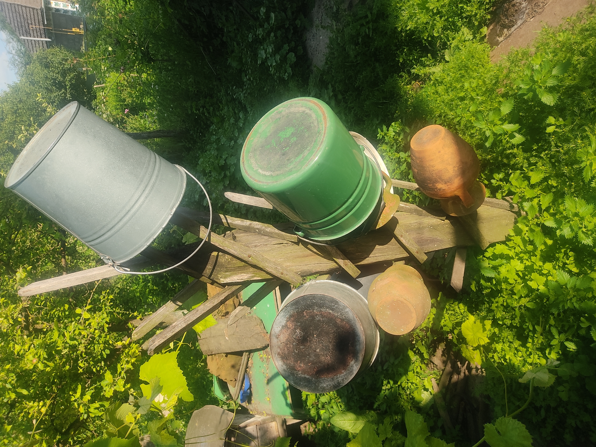 In Voludryntsi, Ukraine, a rural backyard scene depicts a wooden structure supporting various containers. At the top, a large, light gray metal bucket is tilted at an angle. Below it, a green plastic bucket is positioned similarly. Beneath the green bucket, a dark-bottomed metal pot rests on what appears to be a stand. Two clay pots, one a lighter brown and the other a darker reddish-brown, are also visible, one below the metal pot and the other to the right of the structure. The surrounding environment is overgrown with lush green vegetation, including dense ground cover and leafy plants. A building with a tiled roof and a chimney is partially visible in the background, along with trees and foliage. The scene is illuminated by sunlight, casting shadows and highlighting the textures of the objects and vegetation.