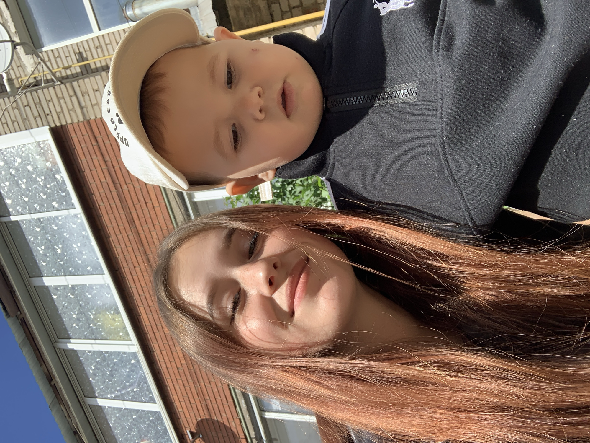 A photo shows a woman and a young child in an outdoor setting. The woman has long, light brown hair and is smiling. The child, wearing a light-colored cap and a black zip-up jacket with a white logo, is positioned above and slightly behind her. The background shows a brick building with windows and a glimpse of blue sky. The photograph appears to have been taken during the day, based on the natural lighting. There are no visible landmarks indicating a specific location in the city of Фоки, Russia.