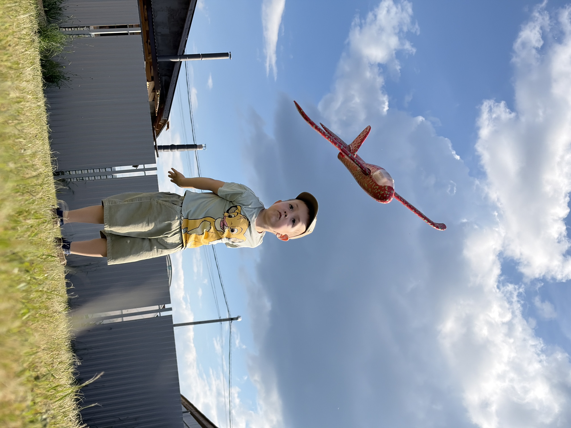 An outdoor, low-angle, full-shot photograph shows a young Caucasian boy, approximately four years old, standing in a grassy yard. The boy is wearing a light green t-shirt with a cartoon lion printed on it, khaki shorts, and a light brown baseball cap. He is looking up and to his left, with his arms slightly extended downwards. A red toy airplane with a white front section is flying in the bright blue sky above and to the left of the boy. The sky is partly cloudy with dark gray storm clouds on the right. In the background, a gray corrugated metal fence runs horizontally across the image. Behind the fence, a building with a dark roof and a chimney is visible on the left, and another building is partially visible on the right. Power lines are strung between poles in the background. The scene appears to be in a residential area, possibly on the outskirts of Chaikovsky, Russia, given the typical rural or suburban setting with fences and residential structures. The lighting suggests it is daytime, with the sun casting shadows.