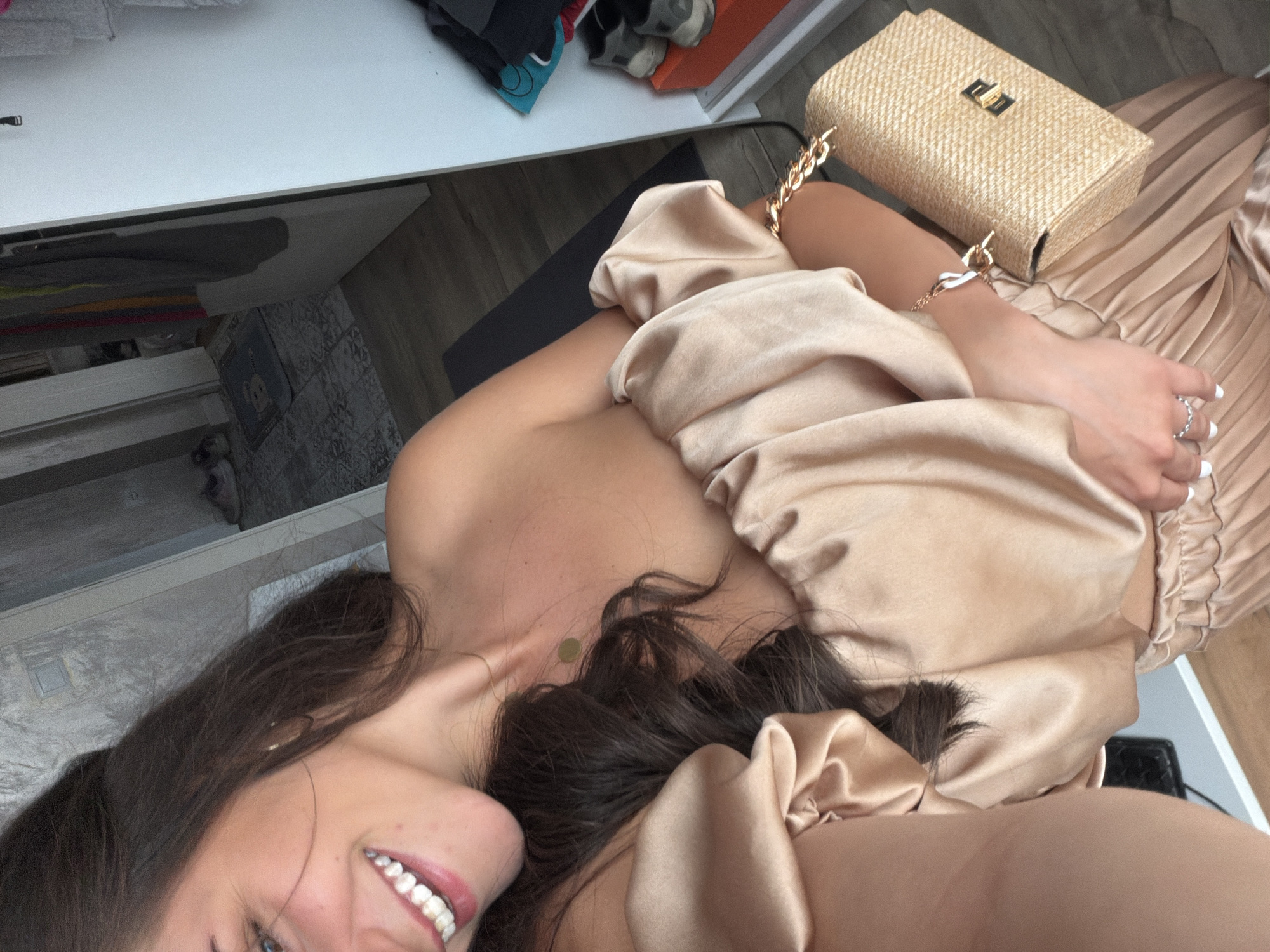 A young woman with long, dark hair smiles at the camera, her head tilted to the side. She is wearing a champagne-colored off-the-shoulder top with puffy sleeves and a matching pleated skirt. A straw clutch with a gold chain strap is held in her right hand, and she wears a silver ring on her left index finger. The background shows an open doorway, a closet with clothing and shoes, and a tiled floor. The location is a residence in Burgas, Bulgaria.