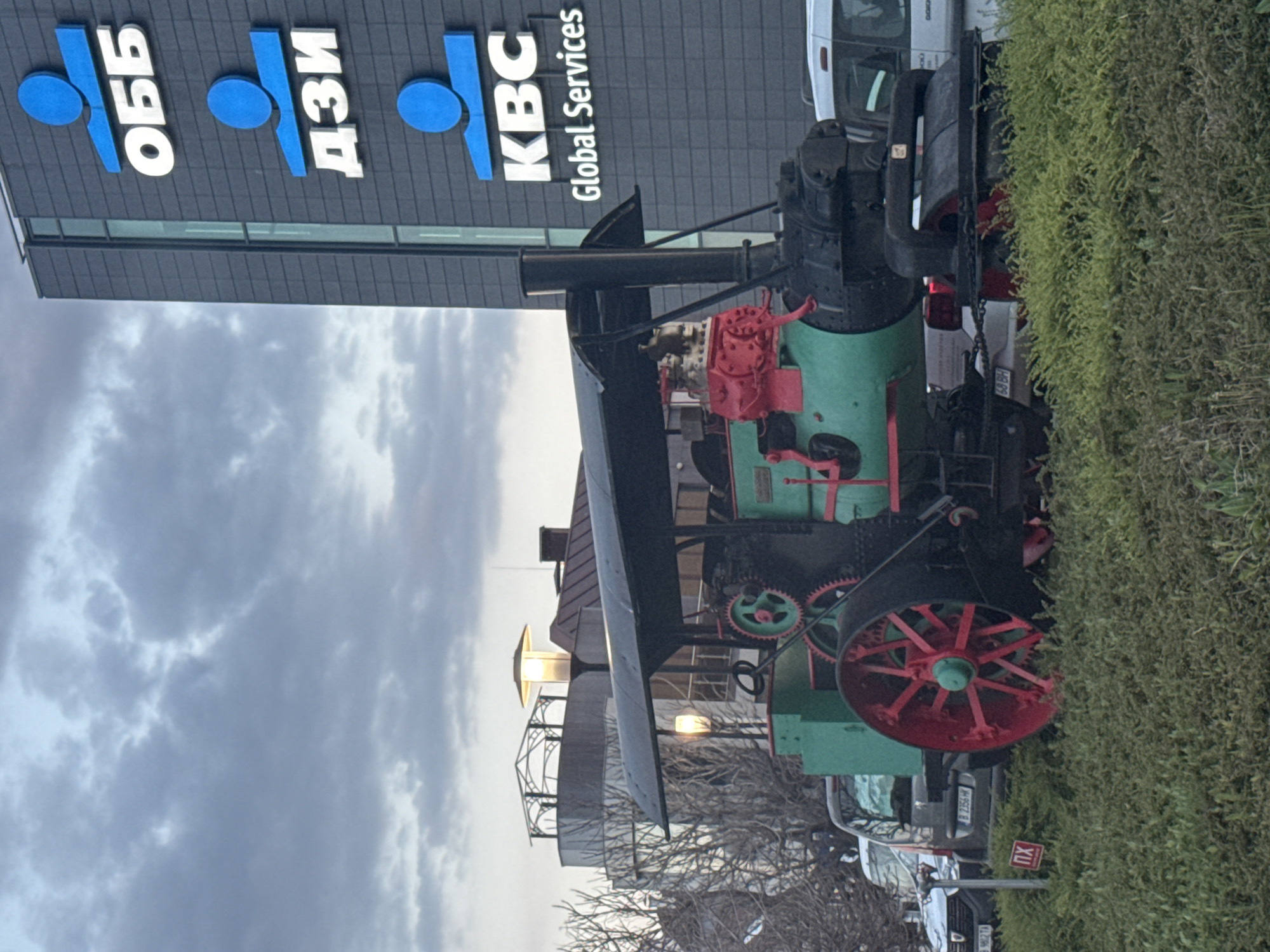 The image shows a vintage, green and black steam tractor, likely a museum piece, situated in front of a modern office building in Varna, Bulgaria. The building has the logos of "OBB," "DZI," and "KBC Global Services" displayed on its facade. The tractor has a red wheel and some red components. A street lamp and bare tree branches are visible near the tractor's front, indicating an outdoor urban setting. A few parked cars are also in view, suggesting the tractor is on display near a parking area or street. The sky is overcast.