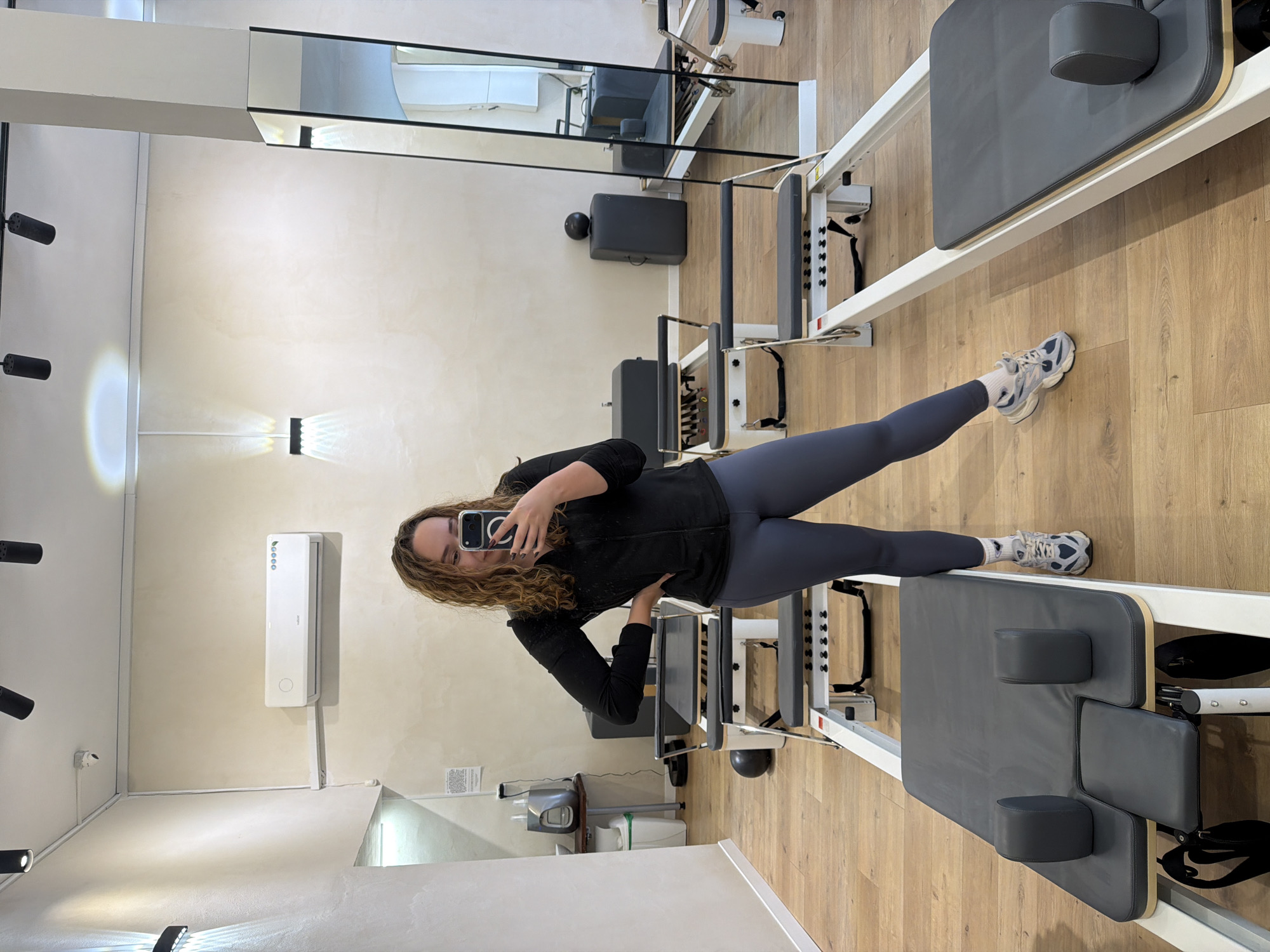 A person is standing in a fitness studio in Ramat Gan, Israel, in front of two Pilates reformer machines. The person is wearing a black jacket, gray leggings, and sneakers, and is holding a phone up to take a selfie. The floor of the studio is wood and the walls are light-colored. There is a mirror on the wall behind the Pilates machines, reflecting the room and equipment. An air conditioning unit and some light fixtures are visible on the wall.