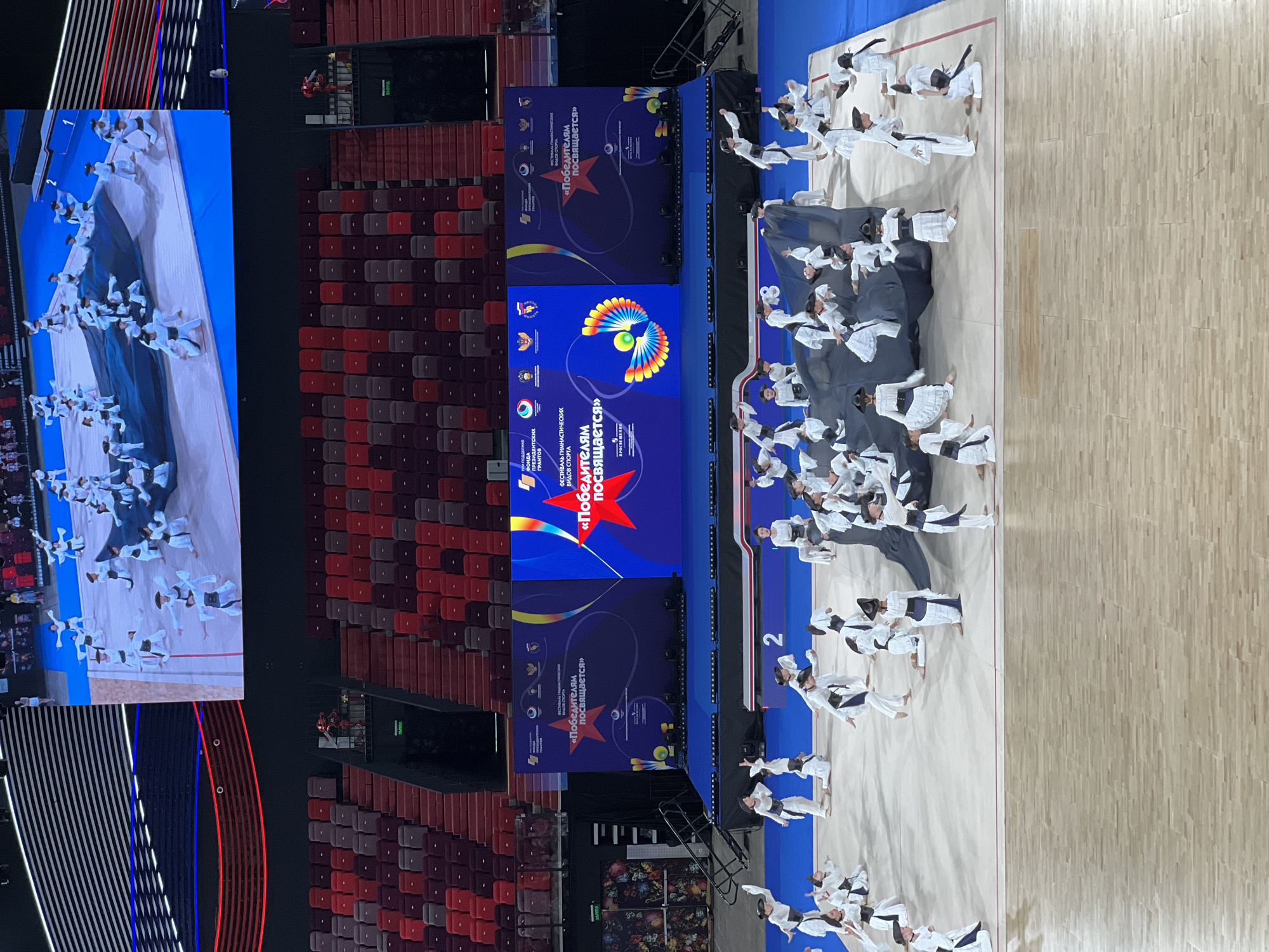 An aerial, wide shot shows a large group of performers in white and black costumes on a light-colored floor in front of a large blue screen. The performers are arranged in a dynamic pattern across the floor, with some kneeling, some standing, and some in motion. A large black fabric is draped across the center of the group. The screen behind them displays graphics and text. In the background, rows of red and burgundy stadium seats are visible, some empty and some occupied by an audience. Above the seats, a patterned ceiling with horizontal lines is illuminated by red and blue lights. The overall scene appears to be a performance or competition held in a large indoor arena.