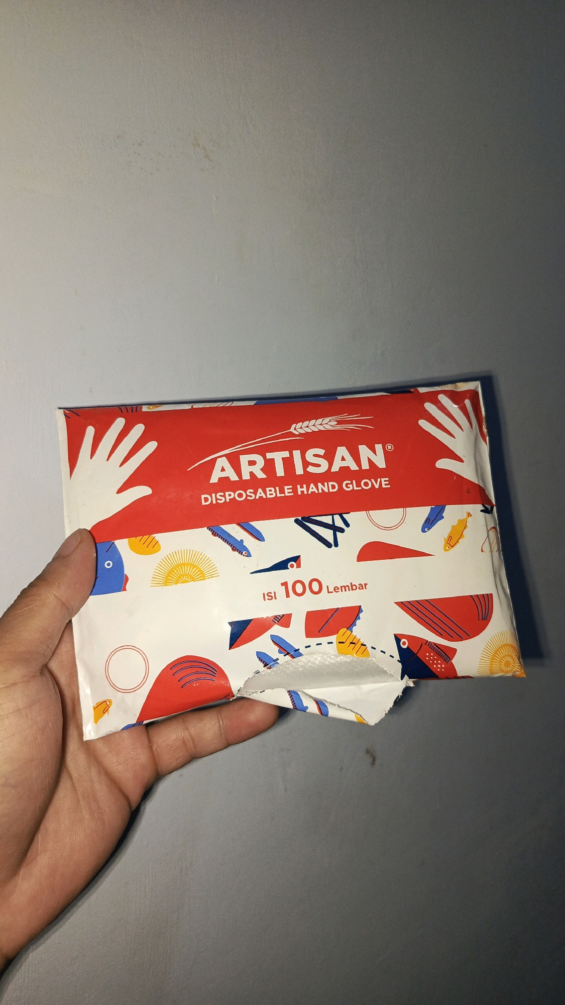A hand is holding a package of Artisan disposable hand gloves against a gray wall. The package features a red band at the top with the "ARTISAN" logo and "DISPOSABLE HAND GLOVE" text. The lower portion of the package is white with colorful illustrations of fish, suns, and other abstract shapes. The packaging is slightly torn open on the right side. There is no indication of the image being taken in Babadan, Indonesia.