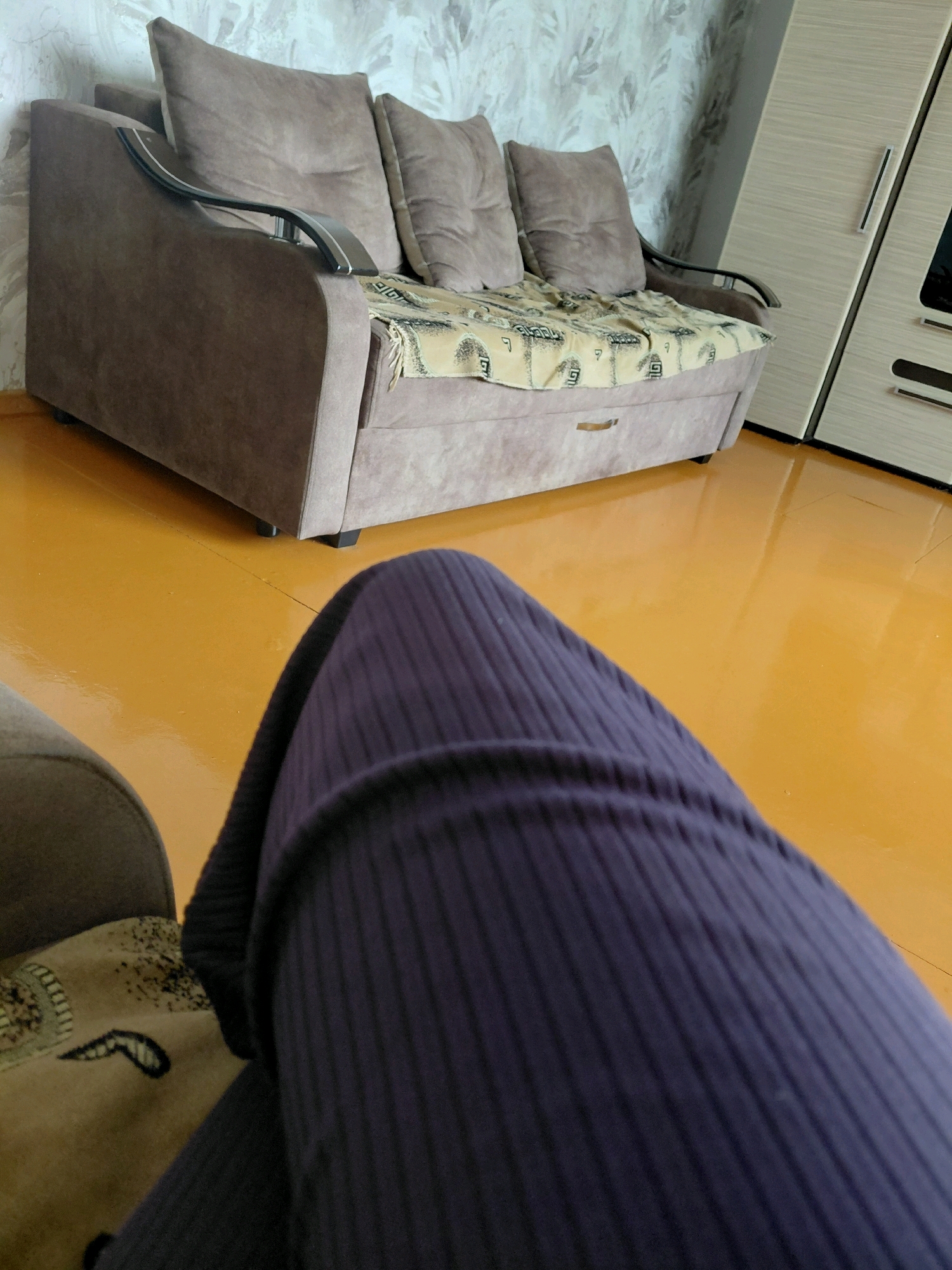 In Malinovka, Russia, an indoor scene depicts a living room with a brown sofa positioned against a wall with textured wallpaper. A beige blanket with a geometric pattern is draped over the sofa. In the foreground, a person's legs are visible, wearing dark purple ribbed leggings. The floor is a glossy, orange-yellow color. To the right of the sofa, a portion of a light-colored wardrobe with horizontal paneling and a dark handle is visible. The scene is illuminated by ambient light.