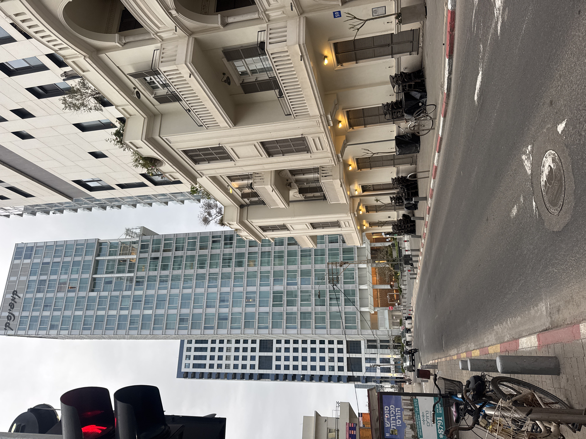 Here is a forensic description of the scene in the image:
**General Impression:** The image depicts a street scene with a mix of architectural styles. A modern high-rise building stands in contrast to older buildings with balconies.
**Location Clues:** The image contains elements suggesting the location is in Tel Aviv, Israel. The Hebrew text on the advertising board and the street sign with the number 59 are indicators of the Middle East. The combination of modern architecture and the old buildings on the other side suggests Tel Aviv, which has a mix of building styles.
**Objects and Details:**
* **Buildings:** On the right, there is a building with balconies. The building is likely an older structure due to its architectural style. A modern building with a grid-like facade stands in contrast to the older buildings. The words "the.fod." are visible on the side of this building.
* **Street:** The street is concrete. There is a red and white painted curb.
* **Traffic Light:** In the foreground, there is a traffic light showing a red light.
* **Bicycle:** A bicycle is visible near the edge of the sidewalk.
* **Advertising:** A sign with Hebrew text is near the bicycle. The text includes words associated with advertising, such as "sales".
* **Chairs:** Stacked chairs covered in black are placed along the side of the street.
**Environment:** The weather looks overcast, which could affect the coloring of the image.
**People:** No people are clearly visible in the image.