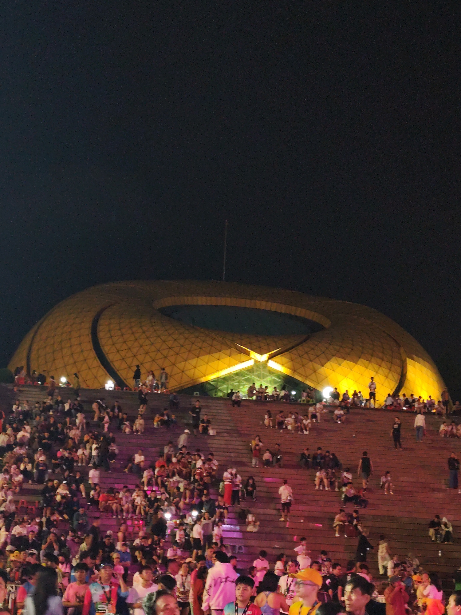 This nighttime photograph captures a large gathering of people on tiered steps leading up to a distinctive, illuminated golden architectural structure identified as the Da Lat Opera House (also known as the Da Lat Flower Garden Conference Center) in Da Lat, Vietnam. The building is donut-shaped, featuring a patterned golden surface brightly illuminated with warm yellow light, and a flagpole is visible above its rear section. A substantial crowd occupies the wide, concrete steps, with individuals standing, sitting, and walking. The density of people is highest in the lower foreground, gradually thinning towards the upper steps and the building. The steps are illuminated with various artificial lights, including white, pink, and purple hues, suggesting an outdoor event. Most individuals are dressed in casual attire.